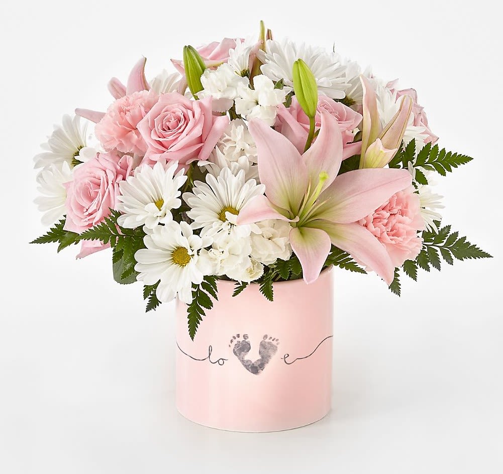 Baby Girl Bouquet - Soft blush roses and pale pink lilies mingle with bright white daisies and carnations in a tender bouquet made for welcoming a new little one.  The mix of pink, white, and fresh green foliage highlights contrasts in texture:  Flowers arrive nestled in a pastel pink cylindrical ceramic printed with a small footprint-and-heart motif, adding a sentimental keepsake touch and tidy presentation that's easy to display.  