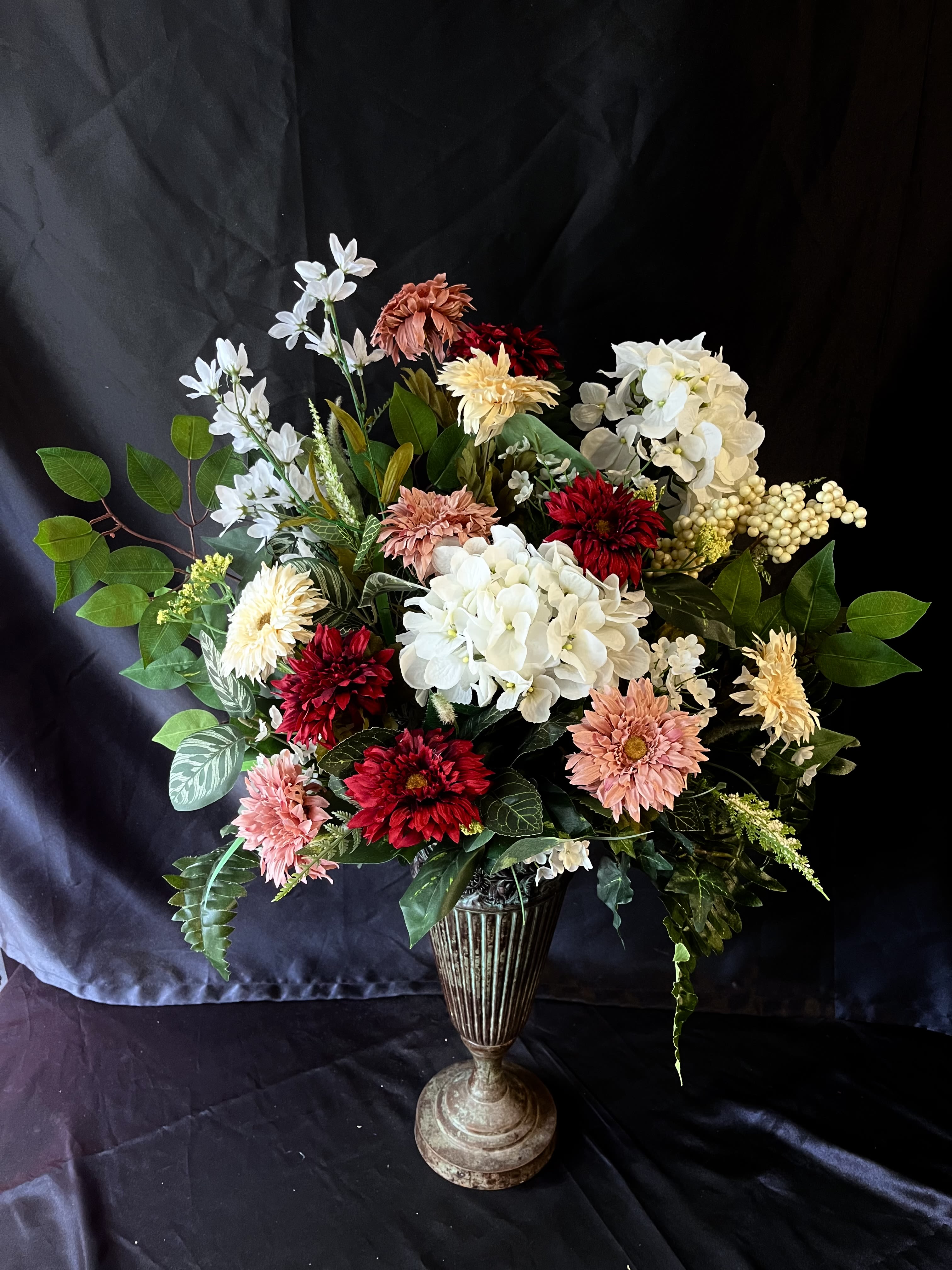 (Silk) Harmony  - A lush cluster of white hydrangea-like blooms punctuated by deep red and soft pink daisies and chrysanthemums creates an immediate, heartfelt impression-perfect as a sympathy arrangement, anniversary bouquet, or a thoughtful everyday gift.  Flowers are layered densely for a full, slightly asymmetrical silhouette: crisp white hydrangea-like heads, red and blush chrysanthemum and daisy-shaped blooms, and pale cream accents woven with broad glossy foliage and a few variegated leaves for contrast and texture. The palette of white, red, pink, and green balances dramatic depth with soft elegance, giving the arrangement visual weight without feeling heavy.  This arrangement is one sided and vase is not included .