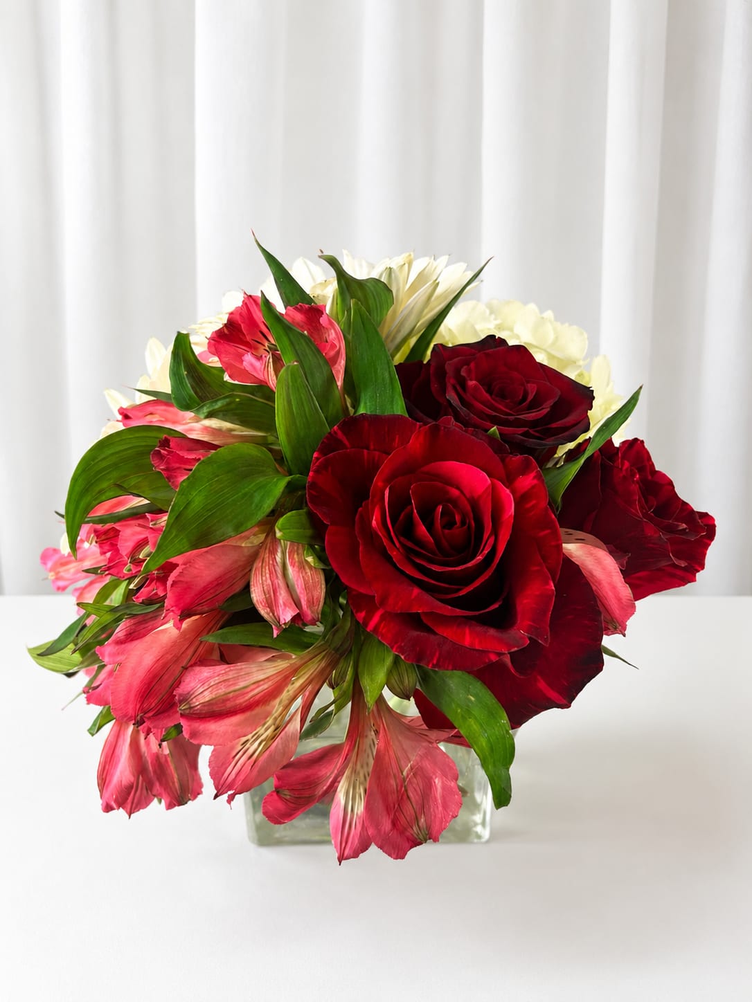 Passion, red - Deep red roses paired with soft pink alstroemeria and pale white blooms create a romantic, celebratory bouquet that reads as a classic mixed arrangement for anniversaries, Valentine's Day, or simply to say "I adore you." The compact design feels intimate and polished-the kind of gift that makes the person you're thinking of feel noticed and cherished.  Flowers are tightly layered in a clear glass vase, where broad green leaves frame the arrangement and give crisp contrast to the rich reds and blush pinks. The dense, rounded shape keeps every stem visible and lush, offering visual depth and a pleasant mix of textures from velvety rose petals to the delicate alstroemeria florets.  Perfect for a bedside table, desk, or small dining centerpiece, this arrangement arrives ready to display in its vase. To keep blooms looking fresh, change the water every two days and trim stems slightly at an angle when you refresh the water.   If any design elements are of major importance to your order, please include them in the florist instructions at checkout or contact us to ensure availability.