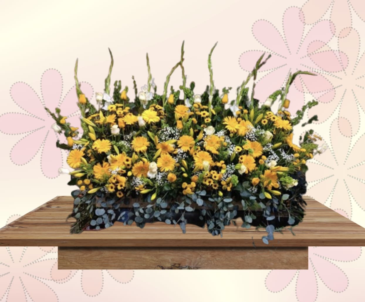 Yellow Sympathy Casket Arrangement - A luminous spray of yellow gerbera daisies and lilies designed for sympathy and remembrance, this casket arrangement brings a hopeful, celebratory touch to a difficult moment. The low, wide profile and generous scale make it ideal for a casket or prominent memorial display, offering a bright, reassuring presence that helps the person you're honoring feel seen and remembered.  Dense layers of sunny yellow blooms are softened by crisp white daisies and lilies, while taller green accent stems give the piece a slightly wild, garden-like silhouette. The composition sits low and broad on a wooden surface in the photo, suggesting a warm, rustic contrast that complements the vivid colors; overall the arrangement reads as full and textured, with a natural, hand-gathered look.  Made to comfort and uplift, this arrangement is a thoughtful way to express sympathy, offer celebration of life, or send condolences to family and friends. Its large size suits funeral services and viewing rooms; please contact us with service details for delivery timing and placement so the tribute arrives prepared for the occasion.