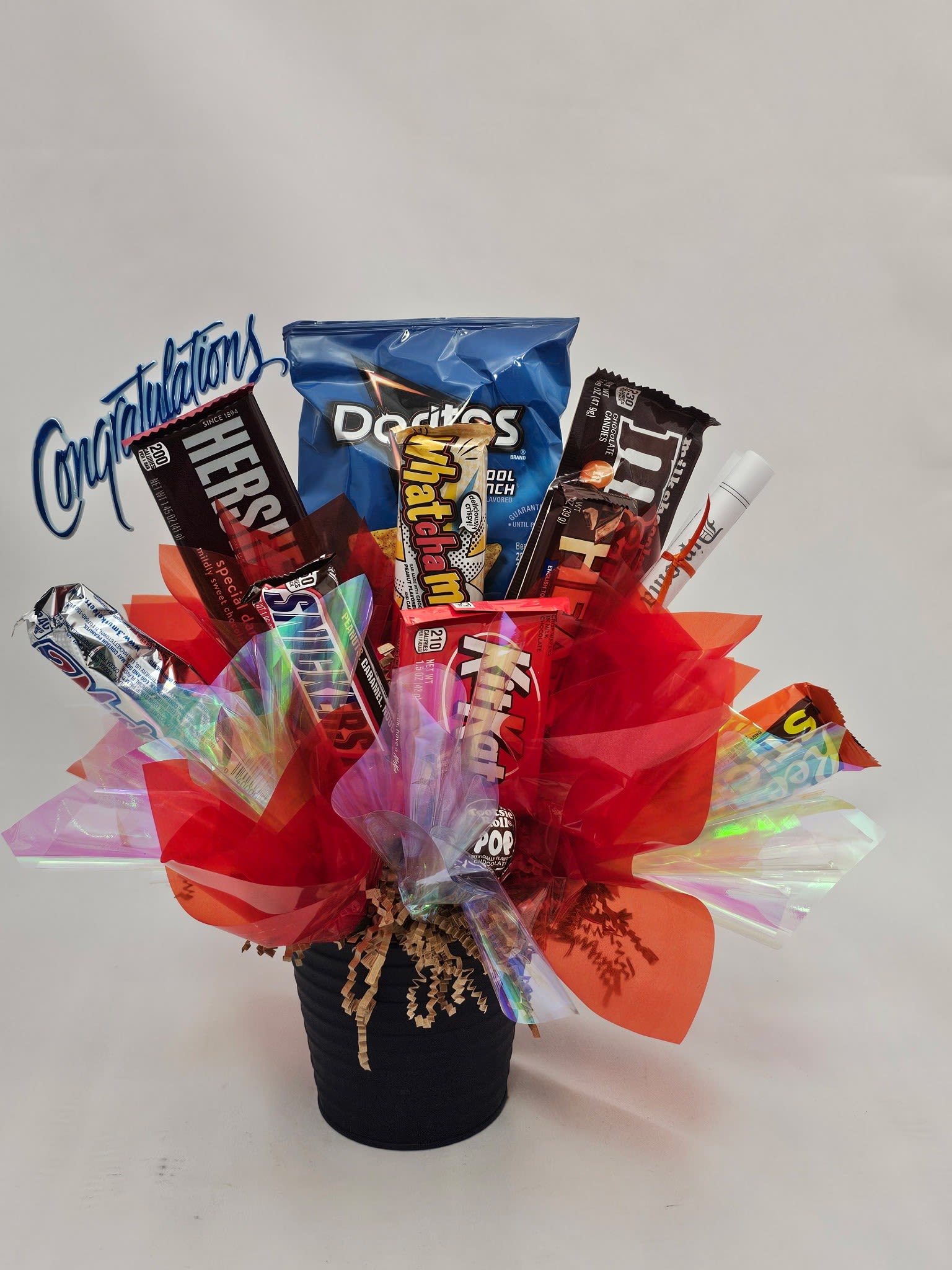 North Montgomery High School Graduation Candy Basket - Bright, playful, and made to celebrate-this graduation candy basket is a cheerful congratulations gift ideal for a North Montgomery High School graduation or any casual celebration. The flower-arrangement-style presentation reads like a festive bouquet, so the person you're celebrating will feel seen and celebrated whether it's for commencement, a birthday, or a job milestone.  Assorted candy bars and snack packs are arranged upright in a sleek black container, with shredded paper filler keeping everything neat and lifted. Layers of red, orange, and iridescent cellophane wrap the base for a lively, colorful pop, while a blue plastic "Congratulations" sign tops the design. Decorative ribbon and clear cellophane finish the basket for a ready-to-gift look.  Perfect for grads, coworkers, or friends who love sweet treats, this is the kind of gift that sparks smiles and lasts through the party. Order in time for ceremony day-no special care required beyond keeping it dry and presenting it unopened for the full visual impact.