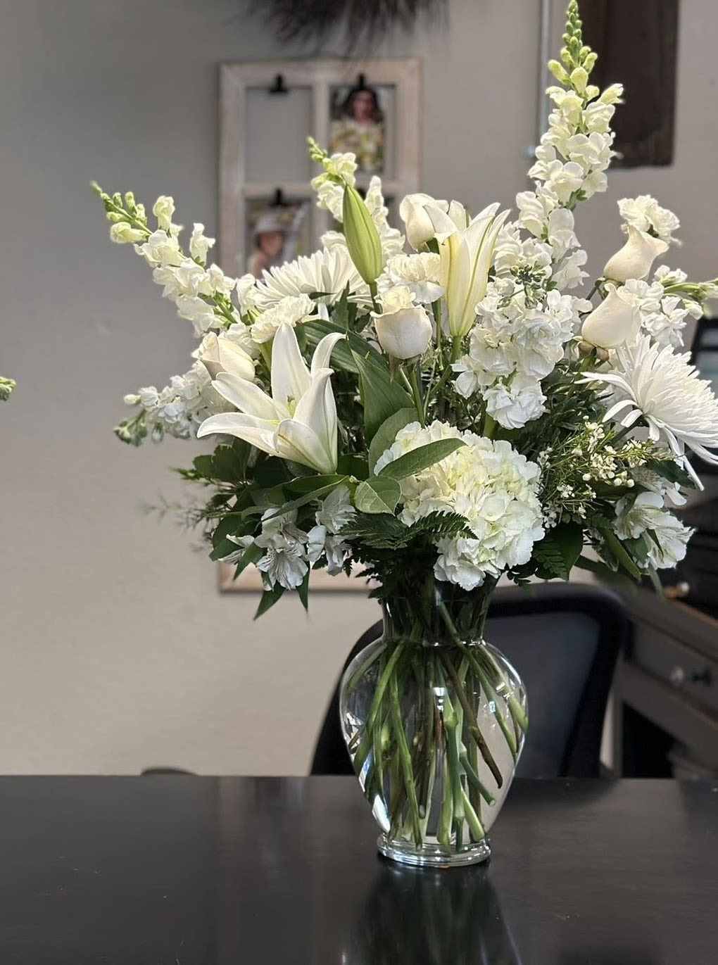 Peaceful elegance in all white  - A serene, all-white bouquet of lilies, snapdragons, hydrangea, and daisy-like blooms or other all white blooms - creates a peaceful focal point that feels both refined and gently expressive - perfect as a sympathy arrangement, a wedding centerpiece, or a thoughtful thank-you gift. The tall, airy design reads calm and fresh; the layered white petals and soft textures send a quiet, comforting message to the person you're thinking of.  Whether you're marking a solemn moment, celebrating a wedding, or simply saying "thank you," this bouquet makes the recipient feel seen and soothed. Delivered ready to enjoy in the clear glass vase, it brings low-maintenance elegance-keep blooms in fresh water and trim stems slightly if desired to extend vase life.