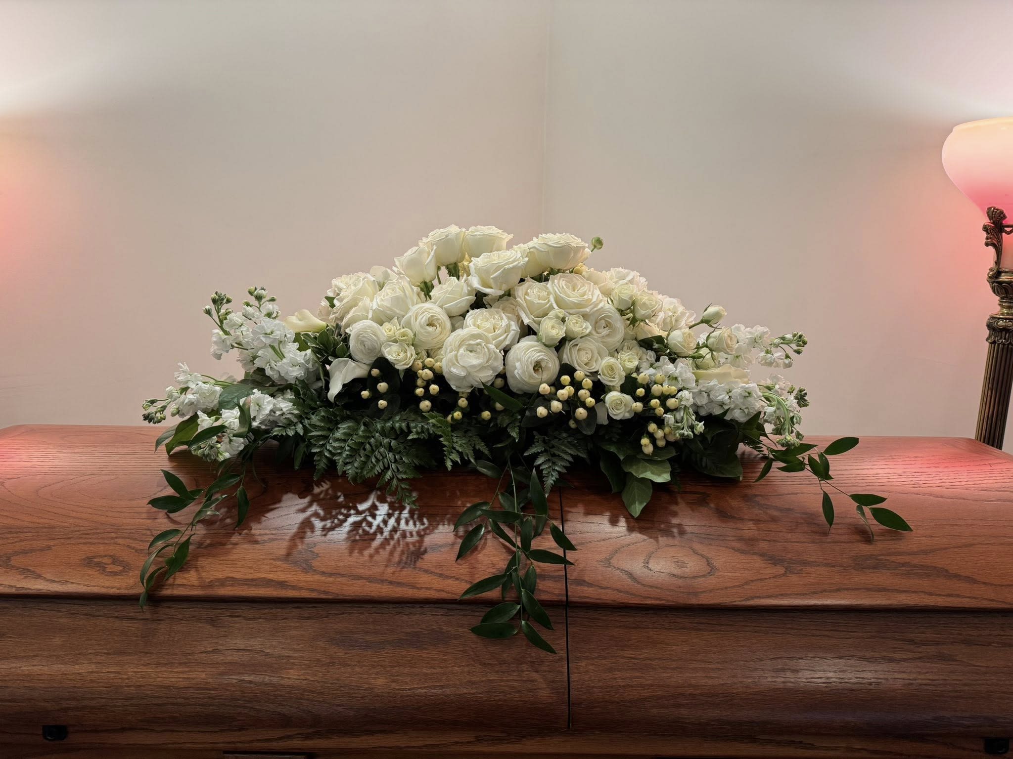 Half casket all white traditional spray  - A serene, low-profile half casket spray in all-white creates a respectful tribute for a funeral or sympathy service. This half casket arrangement of white roses, ranunculus, carnations and like blooms, is designed to convey calm and dignity.   Which is perfect for honoring a loved one with a traditional, understated statement.  The spray is a dense, elongated mound centered across the top of the casket with layered white roses and ranunculus-style petals giving soft, cloud-like texture. Small white filler blooms and clusters add gentle detail while trailing greenery drapes gracefully over the edges, providing subtle movement and a natural finish. The overall palette of white and green keeps the presentation formal and peaceful.  Made to sit low across half of the casket, this arrangement suits family-led services and formal memorials where a classic look is desired. As with all fresh floral tributes, keep the piece cool and out of direct sun until placement; Frazer Flower Shop arranges and delivers with care to ensure the spray arrives ready to honor the person you're remembering.
