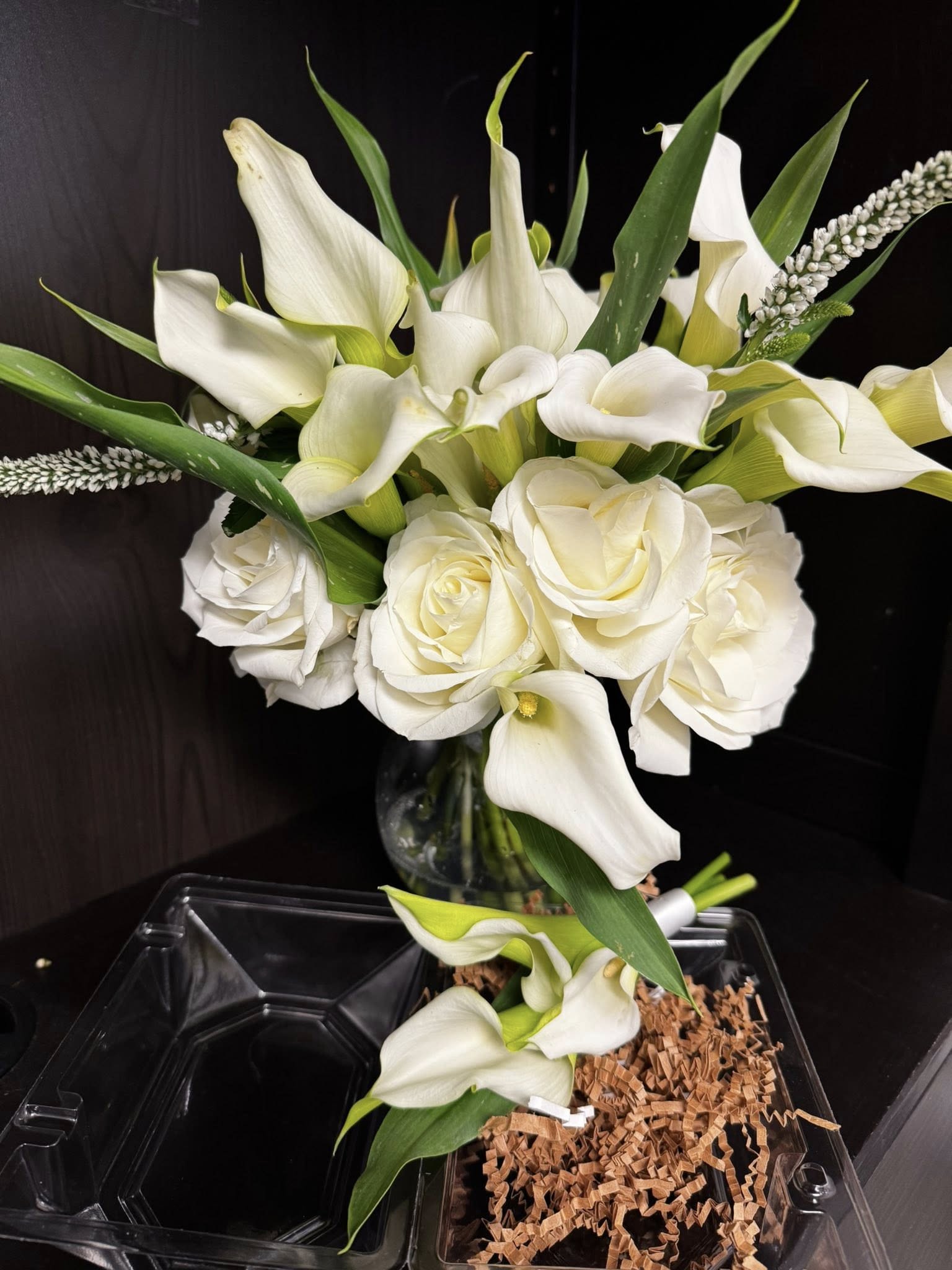 Calla and rose duo - White calla lilies paired with cream-white roses create a serene, refined bouquet perfect for a wedding centerpiece, a sympathy arrangement, or a thoughtful "thinking of you" gift. The rounded, layered composition and soft white-and-green palette give the recipient a calm, elegant impression-ideal for prom corsages, memorial services, or any moment when you want someone to feel quietly comforted and seen.  This arrangement features sculptural calla lilies and velvety cream roses arranged with long green leaves and delicate white textured accents for subtle height and contrast. Presented in a clear glass vase set on a clear plastic tray with brown shredded paper filler, the design balances formality with simplicity so it complements both modern and traditional settings. The compact, rounded shape makes it easy to place on a table or mantel; care is simple-keep water fresh and trim stems as needed to maintain its graceful silhouette.