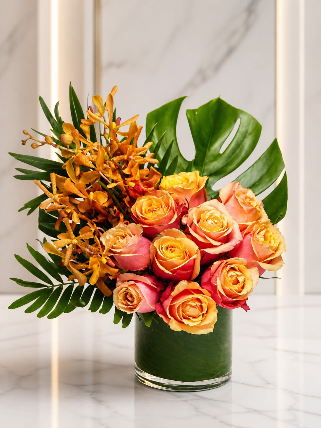 Sunset Mirage - A bold tropical-inspired arrangement designed with radiant sunset tones and lush modern textures. Sunset Mirage features premium free spirit roses in warm peach, coral, and golden sunset shades accented with vibrant mokara orchids and sculptural tropical greenery for a rich, contemporary presentation. Arranged in a modern leaf-lined clear cylinder vase, this design brings together elegance and tropical warmth in a luxurious all-around composition perfect for birthdays, celebrations, thank-you gifts, or statement floral décor.  To ensure the freshest presentation possible, certain flowers or greenery may occasionally be substituted with blooms of equal or greater value while maintaining the overall tropical color palette and modern design style. Similar premium roses, orchids, tropical foliage, or accent blooms may be used based on seasonal availability. The container may also vary slightly depending on inventory, but the arrangement will always be created in a comparable modern premium vase that complements the overall aesthetic and value of the design.