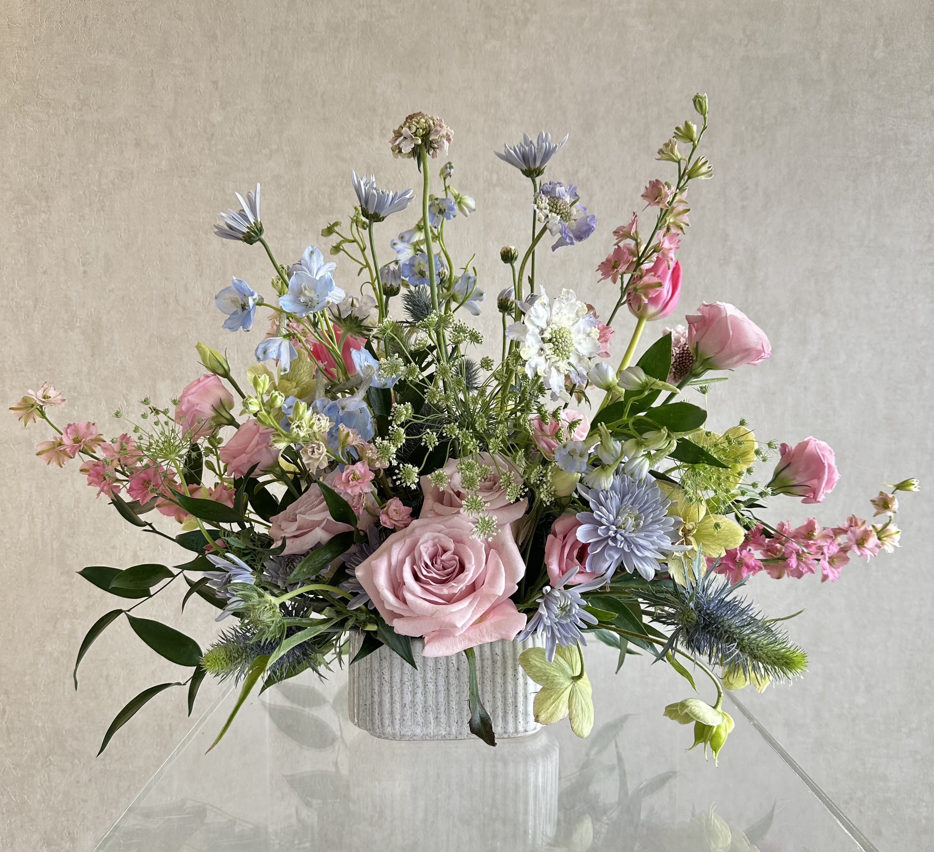 Ocean Whisper - A soft, garden-style arrangement is a pastel flower arrangement perfect for birthdays, spring celebrations, or a thoughtful thank-you. The design centers on a large blush rose surrounded by airy stems of delphinium, mums and other clustered blooms in pink and pale blue, white and fresh green. Its light, whimsical silhouette makes the recipient feel noticed and gently celebrated beautifully set in a ribbed white ceramic vase