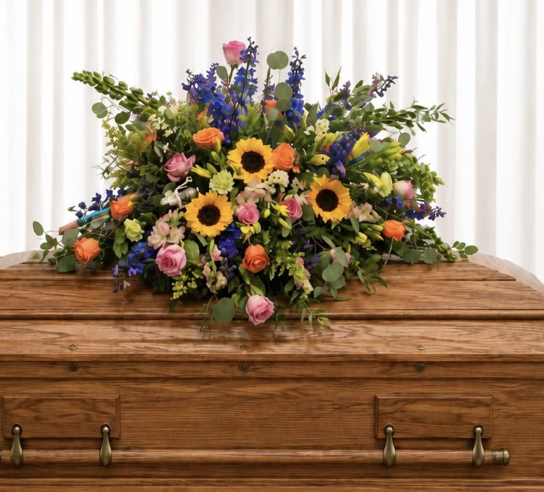 Spring fields - Bright, garden-fresh and abundantly sympathy spray. Sunflowers and coral-pink roses create a focal point while tall blue delphinium add elegant height and a striking cool contrast.  The design is dense and layered in a garden-style composition: bright yellow sunflowers, pink and coral roses, blue delphinium, alstroemeria, snapdragons and mixed seasonal blooms woven with airy greenery. It's a sprawling centerpiece with plenty of textural variety-round, velvety roses against spiky delphinium and sculptural snapdragons-so it reads full and abundant from every angle. The vivid mix of yellow, pink and purple tones keeps the arrangement lively and eye-catching.  someone who loves bold color and lush, garden-inspired arrangements; order with confidence knowing this arrangement was composed to look full and fresh on delivery.   If any design elements are of major importance to your order, please include them in the florist instructions at checkout or contact us to ensure availability.