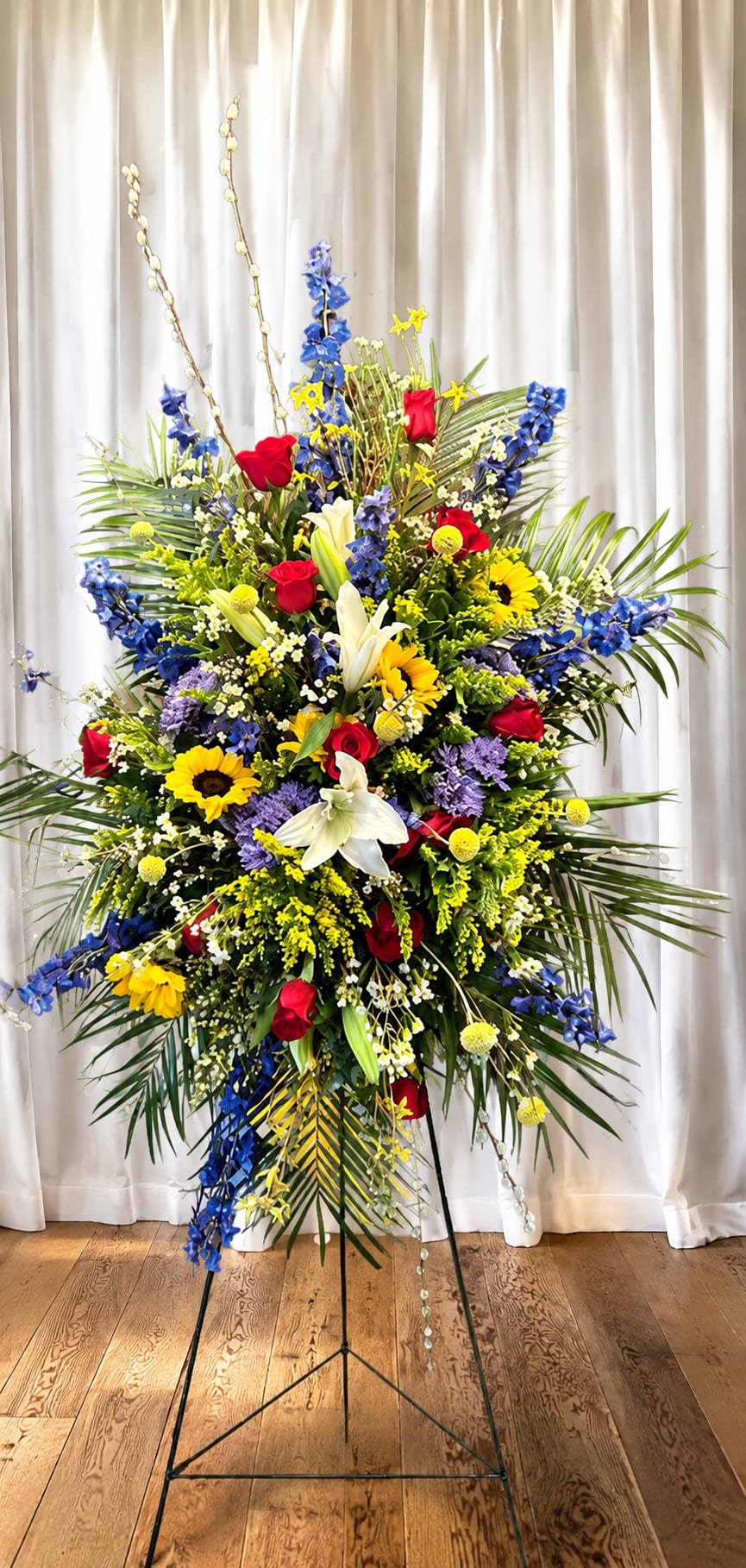 Spring memories - Bright, high-contrast blooms make a lasting impression with this large standing spray for funeral or memorial services. The vertical profile and metal easel stand present red roses, white lilies, yellow sunflowers and vivid blue flowers in a dense, layered composition that reads both celebratory and reverent-the kind of arrangement that honors a life while bringing color and warmth to a service.  Rich texture comes from long palm fronds and mixed filler blossoms that lift and frame the focal flowers, creating dramatic height and depth. The palette of red, white, blue and sunny yellow provides a strong visual tribute suitable for formal settings; the arrangement's scale and easel make it ideal for placing near a casket, podium, or memorial table. Designed to arrive ready-to-display, it conveys sympathy and remembrance while celebrating a vibrant life.   If any design elements are of major importance to your order, please include them in the florist instructions at checkout or contact us to ensure availability.