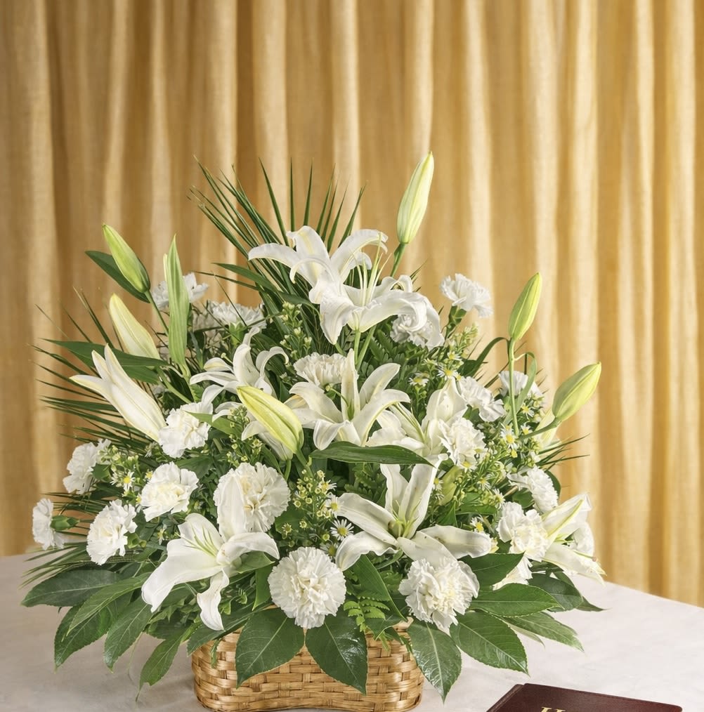 Majestic white - Pure white lilies and soft white carnations form the heart of this Majestic white flower arrangement, a calm and elegant choice for sympathy, a thoughtful thank-you, or a general gift to let someone know you're thinking of them. The low woven basket gives the design a grounded, classic feel while unopened lily buds lift the composition with a fresh, just-opening promise-perfect for conveying quiet comfort or gentle celebration to the person you have in mind.  A full, rounded design pairs luminous white blooms with small daisy-like filler flowers and dense green foliage for contrast and texture. The yellow centers of the daisies add a subtle warm note that keeps the palette from feeling austere. Arranged in a sturdy woven basket, this piece sits low and wide-making it an ideal centerpiece for a table or a peaceful addition to a bedside or mantel.  Choose this arrangement when you want to make someone feel truly seen with a serene, refined floral gift; it's especially fitting for moments that call for graceful restraint. The basket construction keeps stems neatly arranged and stable-no extra vase needed-so it's ready to display on arrival and easy to care for with regular water refreshes.   If any design elements are of major importance to your order, please include them in the florist instructions at checkout or contact us to ensure availability.