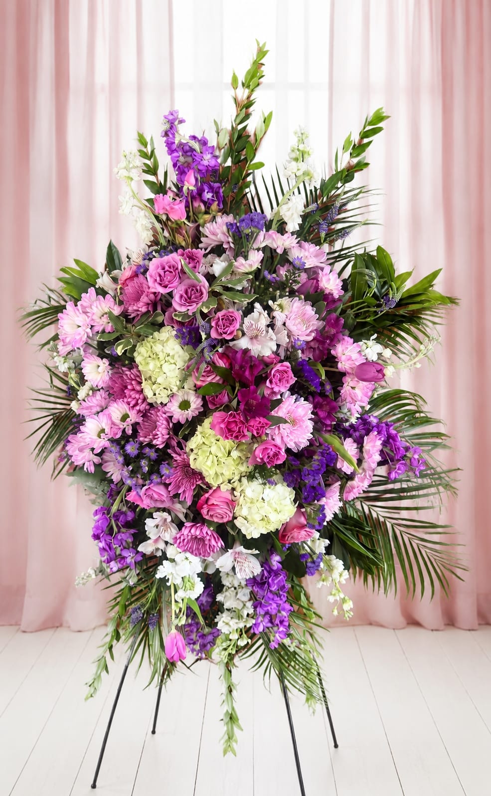 Whisper of Grace - A lush, formal standing spray that reads as both a tender sympathy tribute and a graceful celebration-perfect as a memorial arrangement or for a ceremonial floral display. Whisper of Grace is a large flower arrangement designed to stand tall on a black metal stand or standing easel, its soft pastel palette of pinks, purples, whites, and pale greens creating an immediate sense of calm and comfort for the person you're honoring.  This composition layers rounded roses, hydrangeas, and chrysanthemum blooms with alstroemeria, daisies, and tall snapdragon spikes for vertical movement; touches of dendrobium orchids add an elegant, airy note. Arching palm fronds and varied greenery give the spray a full, sculpted silhouette with deeper violet accents punctuating the softer tones. The result is dense and ceremonial without feeling heavy-balanced, textured, and visible from a distance.  Made to convey sympathy, respect, or a heartfelt remembrance, Whisper of Grace offers a meaningful way to show you're thinking of someone during a service or memorial gathering; it also suits dedications and formal celebrations where a dignified floral presence is desired. Arrives ready to display on the included stand or easel-no assembly required-and, like most mixed fresh arrangements.  If any design elements are of major importance to your order, please include them in the florist instructions at checkout or contact us to ensure availability.