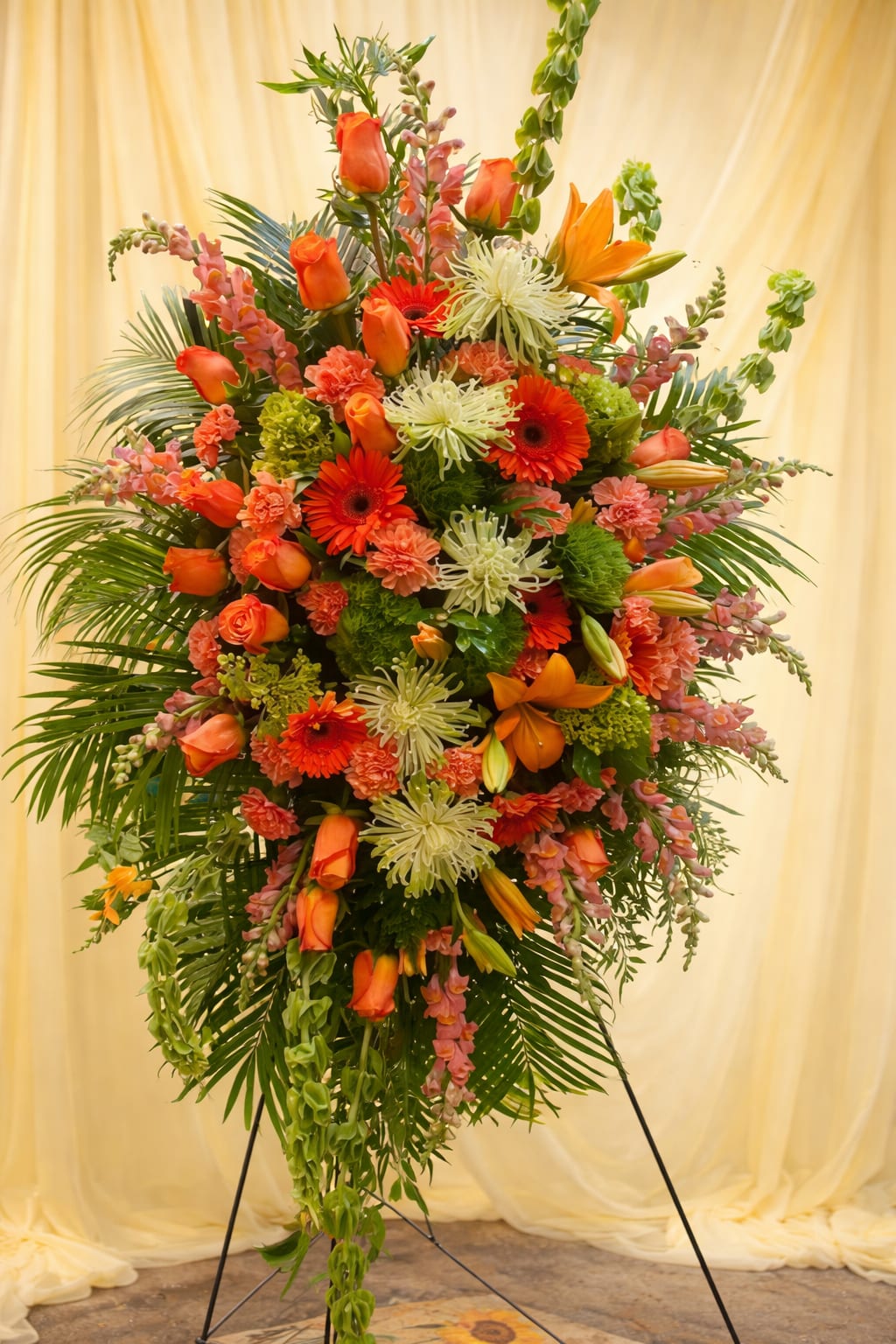 Orange beauty  - A tall, layered standing spray in warm orange and soft pink-this funeral or sympathy arrangement makes a respectful, uplifting statement for a memorial service. The vertical design on a metal easel presents orange roses, orange gerbera daisies, and orange lilies as the visual anchors, while pink carnations and white spider chrysanthemums add gentle contrast and a comforting softness the person you're remembering will be honored by.  The composition is dense and formal, with blooms arranged around a central core and flowers extending outward and downward to create graceful movement. Abundant greenery frames the flowers, enhancing the bright but reverent palette of orange, pink, green, and white. Mounted on a sturdy standing easel, it's sized to be clearly visible beside a casket or at the front of a service and designed to hold its shape throughout the ceremony.  Choose this arrangement when you want to convey warm remembrance and heartfelt sympathy-the colors feel uplifting without being celebratory, and the tall presentation ensures the tribute reads with dignity. Care and delivery are arranged to keep the flowers fresh for the service; please confirm easel delivery requirements and service timing when you place your order so the memorial receives the tribute at the right moment.   If any design elements are of major importance to your order, please include them in the florist instructions at checkout or contact us to ensure availability.  