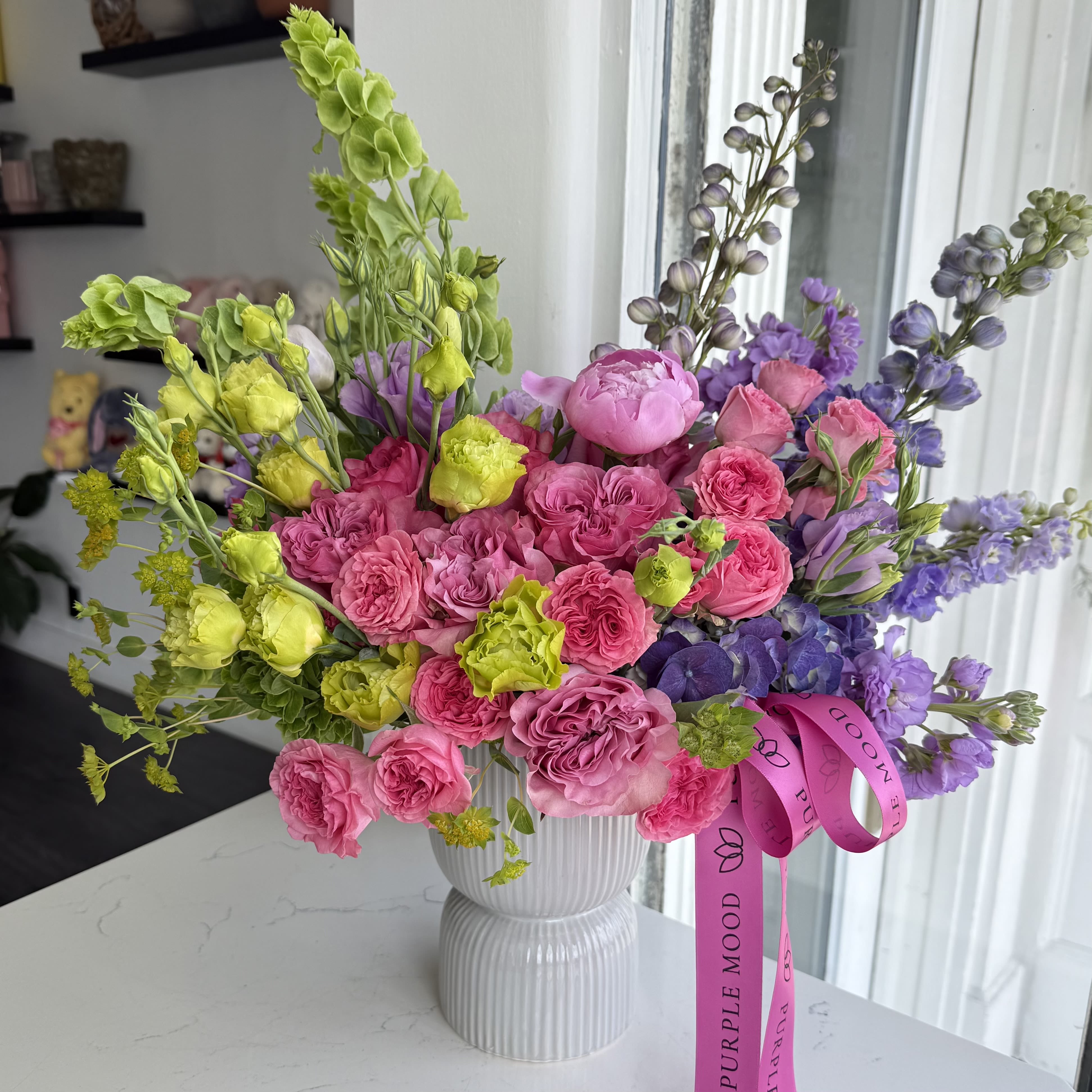 Sweet Secret Vase - A vibrant garden-style arrangement featuring lush pink garden roses, delicate lavender blooms, and fresh seasonal accents in soft greens and purples. Designed with natural movement and texture, this elegant piece brings a romantic and joyful spring feel to any space. Arranged in a modern ceramic vase and finished with Purple Mood signature ribbon. Vase included.
