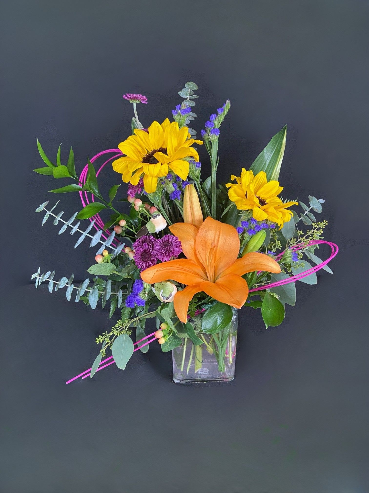 Bright mixed bouquet with sunflowers, orange lily, and purple accents in a glass vase