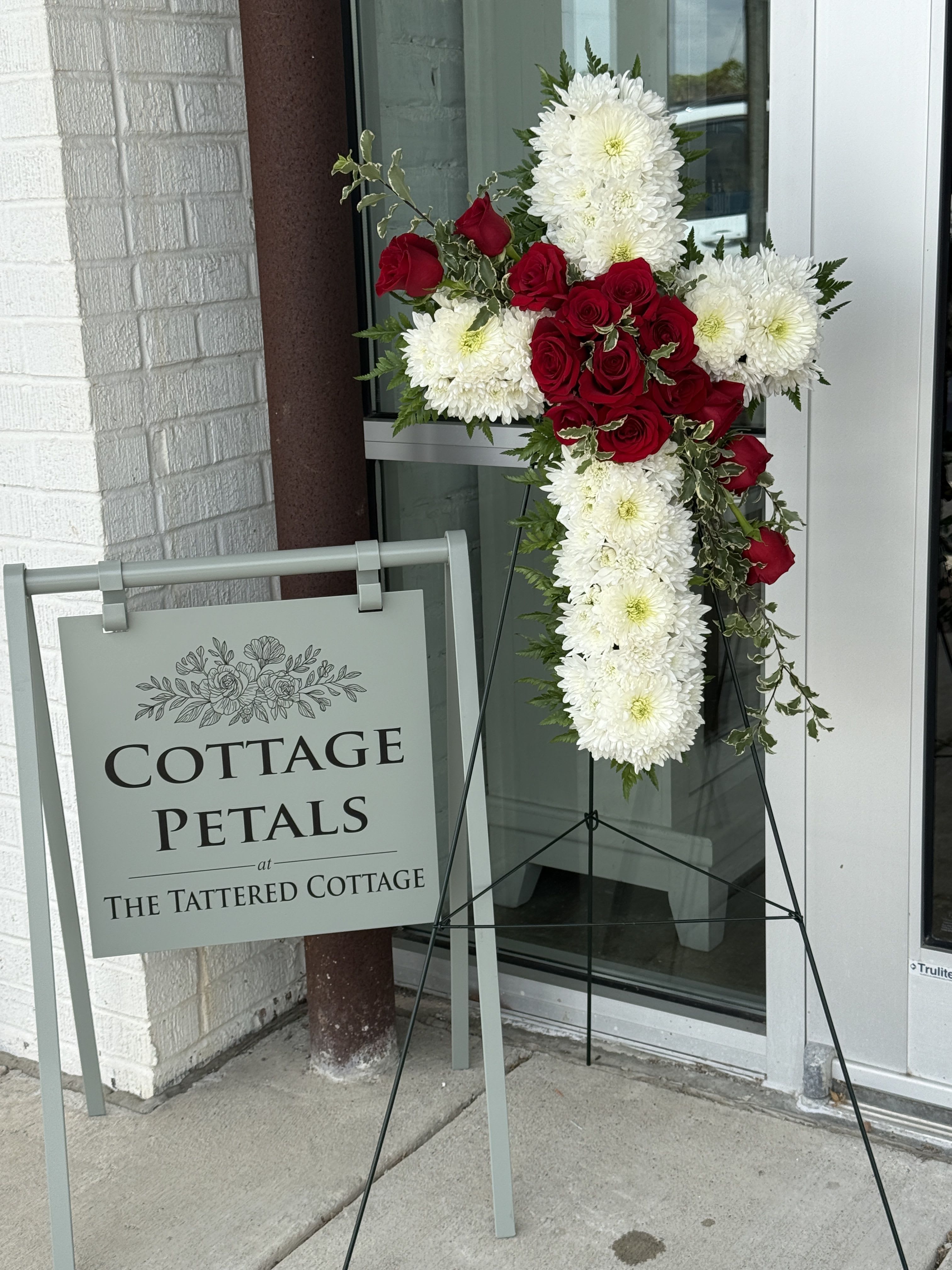 Crimson Grace Cross - A beautiful and meaningful expression of love, faith, and remembrance, the Crimson Grace Cross is designed with rich red roses and soft white chrysanthemums arranged in the shape of a cross. Accented with flowing greenery and natural texture, this standing tribute offers comfort and honor while celebrating a life filled with love and cherished memories.  The elegant contrast of deep red and crisp white creates a timeless design suitable for funeral services, memorials, and celebrations of life.  Flower varieties and greenery may vary slightly based on seasonal availability, but the overall style, fullness, and color palette will always be maintained.