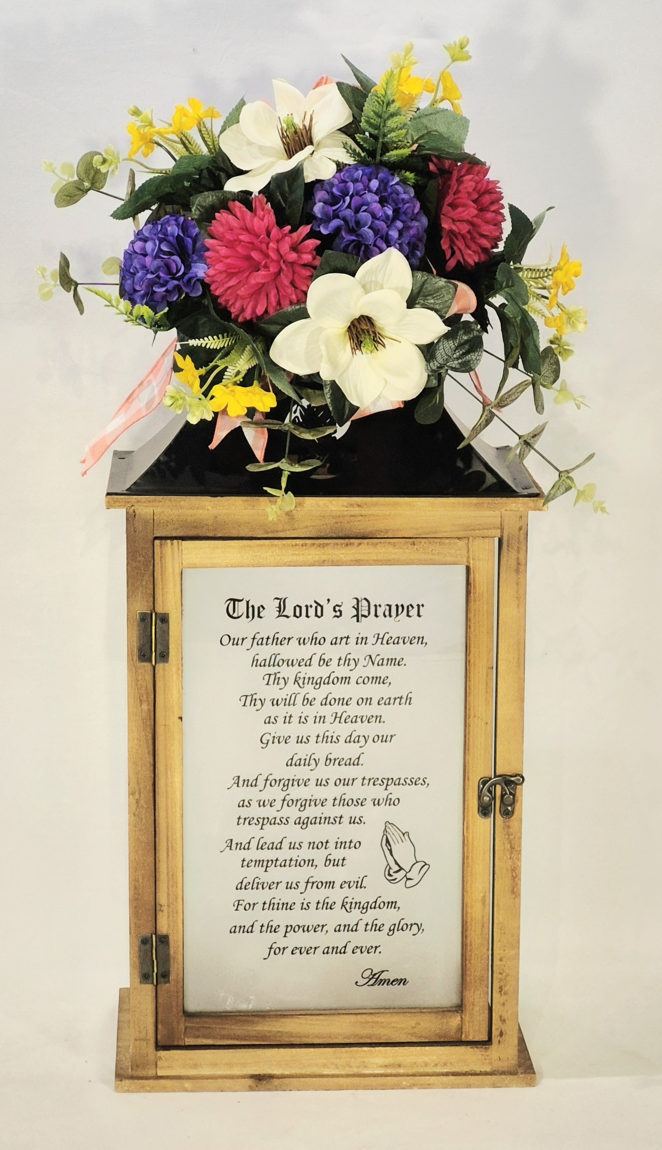 Lord's Prayer Wooden Lighted Lantern  - A softly luminous memorial piece, this flower arrangement with a Lord's Prayer wooden lighted lantern combines devotional sentiment with decorative charm-perfect for a sympathy display, church gathering, or a thoughtful remembrance. The wooden cabinet features a printed prayer panel behind a glass front, giving the piece a reverent focal point while the surrounding florals speak comfort and warmth to the person you're honoring.  Complementary silk blooms in white, bright pink, purple and small yellow accents form a dense, rounded bouquet that sits atop the lantern; dark green foliage and a few trailing stems frame the composition and add gentle texture. The glass-fronted cabinet and prayer panel lend a quiet, sacred quality while the colors bring an uplifting visual note to the arrangement.  Sized to sit on a memorial table or at the front of a church service, the lantern arrangement stands 27" tall, 11" wide and 8" deep-large enough to be a meaningful tribute without overwhelming a room. The silk flowers require no water, making this a long-lasting keepsake that will continue to comfort the recipient and honor the memory for weeks to come.