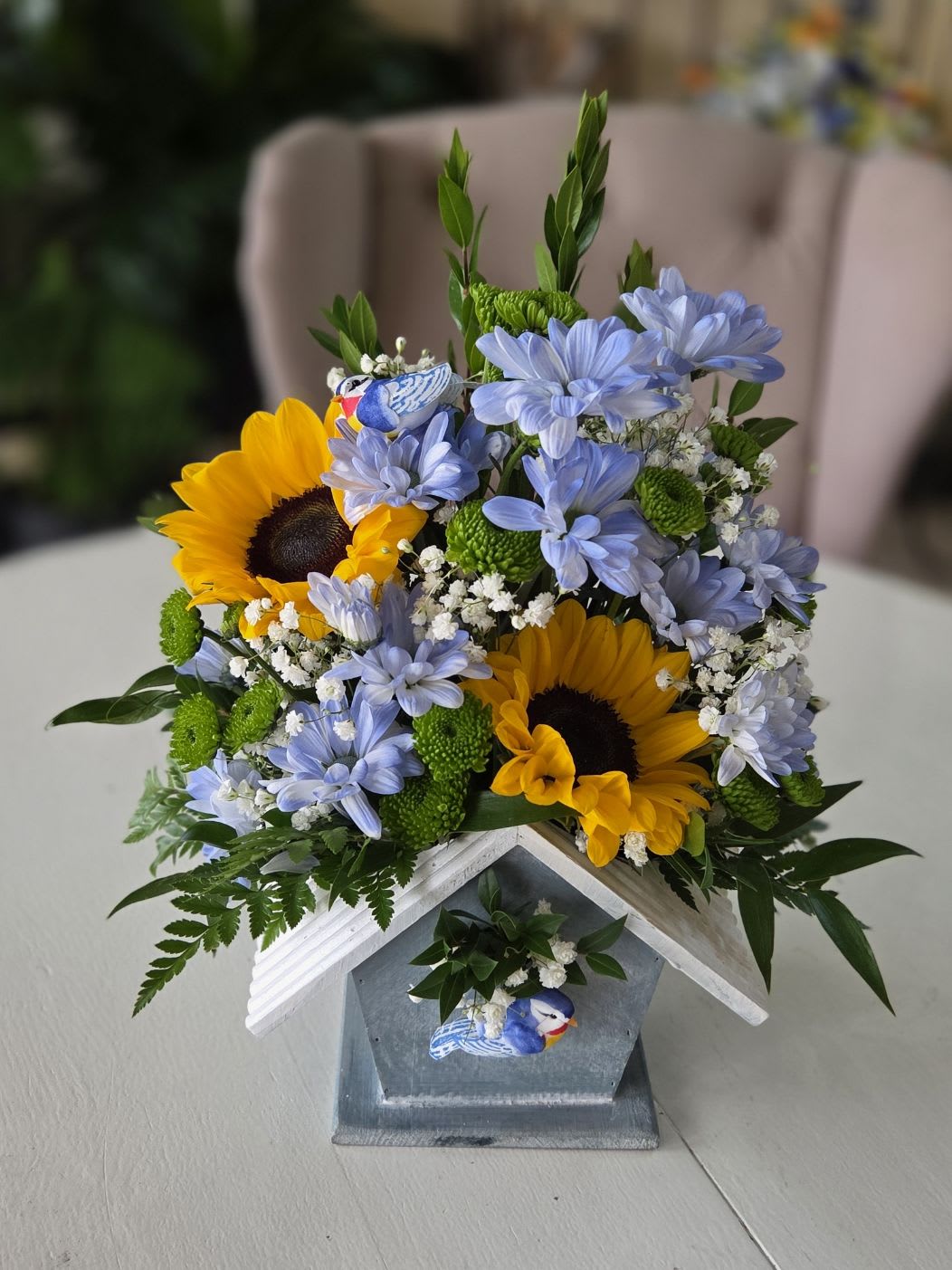 Sunny Songbird Cottage - A cheerful and charming floral arrangement featuring radiant sunflowers, soft lavender daisies, green button mums, and delicate baby’s breath nestled inside a rustic decorative birdhouse container. Accented with whimsical blue bird details and lush greenery, this design brings warmth, happiness, and a touch of countryside charm to any space. Perfect for birthdays, get well wishes, spring celebrations, or simply brightening someone’s day.