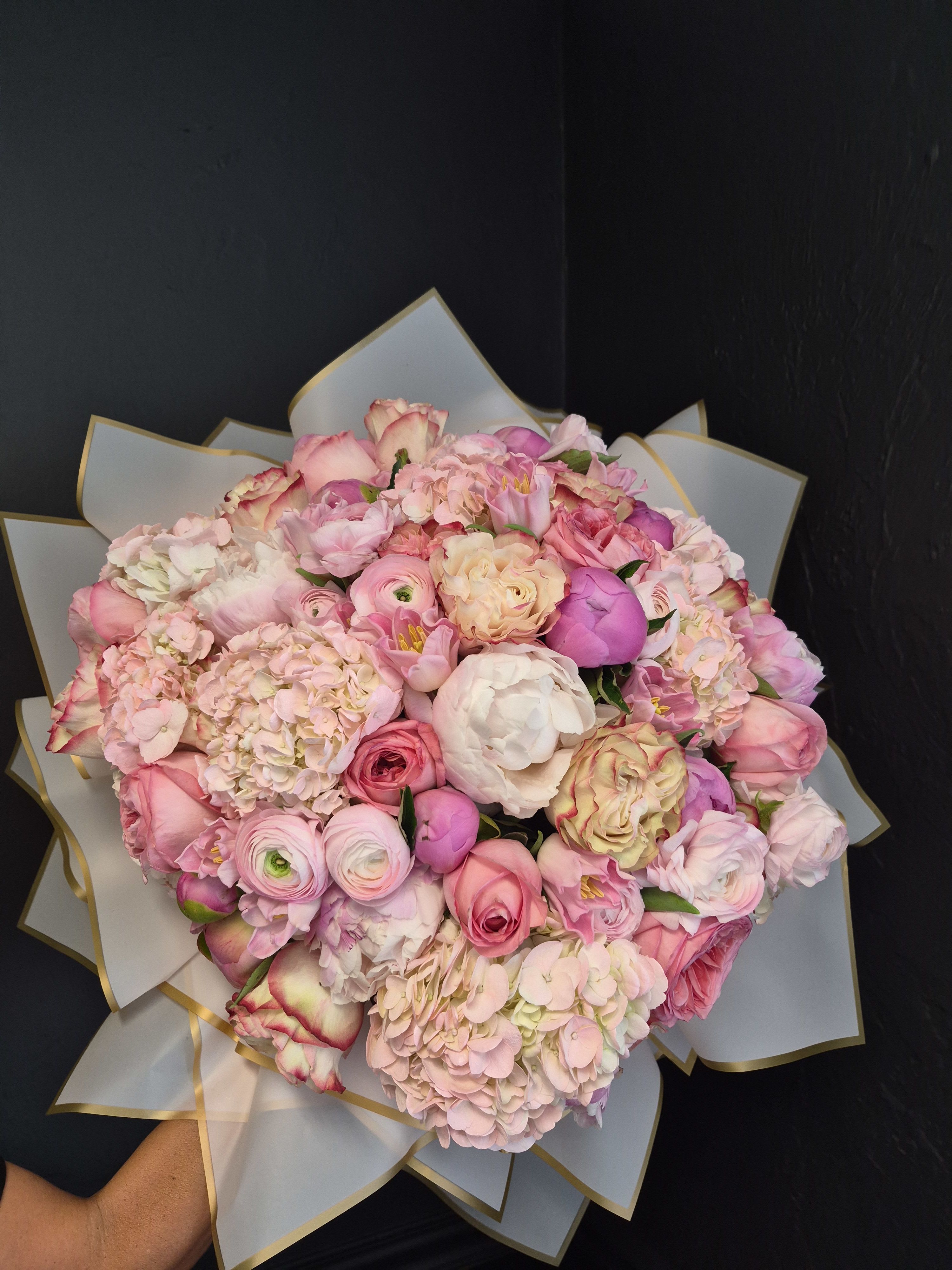 Blooming Abundance - Blooming Abundance is a lush, hand-tied bouquet overflowing with softness, romance, and garden-inspired beauty. Designed with layers of premium seasonal blooms in delicate shades of blush pink, soft ivory, peach, and lavender, this arrangement evokes a feeling of femininity, tenderness, and quiet luxury.  Voluminous hydrangeas create a cloud-like foundation, while peonies, garden roses, ranunculus, and tulips unfold naturally throughout the bouquet, adding rich texture, movement, and depth. The rounded, abundant silhouette feels both timeless and expressive — like a flourishing garden gathered lovingly by hand.  Created for meaningful celebrations and heartfelt moments, Blooming Abundance is especially beautiful as a bridal bouquet, anniversary gift, birthday surprise, or an offering of love and appreciation. Beyond their beauty, flowers have a way of bringing warmth, softness, and emotional presence into a space, helping people feel cherished, uplifted, and deeply seen.  Designed in our signature romantic garden style, this bouquet is meant to make an unforgettable impression the moment it arrives. For best longevity, place in fresh water promptly and recut stems before arranging in a vase.