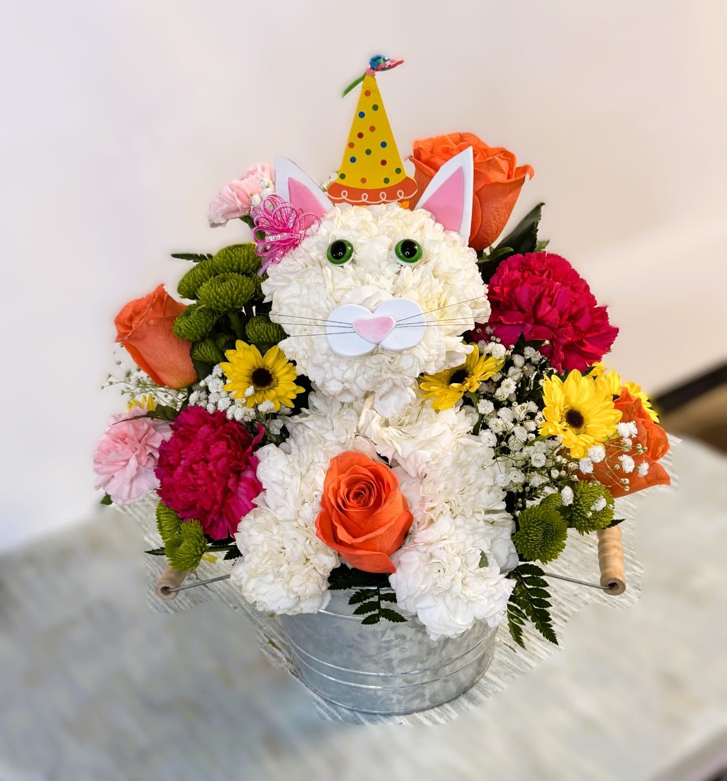 Birthday #11 - A playful, pet-themed birthday flower arrangement that makes the recipient feel delightfully celebrated: a whimsical cat  sculpted from white carnations with plastic eyes and nose, framed by bright orange roses, hot-pink and magenta carnations, cheerful yellow daisy-style blooms, purple filler flowers, green button mums, and clusters of tiny white accents. Perfect as a birthday bouquet, congratulations gift, or an anytime pick-me-up for someone who loves animals and color, it reads as joyful and childlike-she'll feel seen and smiled at the moment it arrives.  The flowers are arranged in a white ceramic container decorated with vertical rainbow stripes and gold starburst motifs and finished with a light lavender ribbon bow and playful hard-goods: a party hat, decorative ears, a decorative cat-face cutout on some variations, a wire handle or metal bucket option, and small plastic dog eye and nose picks that give the sculpted face its animated expression. Compact and densely packed (measuring about 5"H x 8.6"L x 4.25"W), this keepsake-style piece ships ready to display; care is simple-keep in fresh water, away from direct heat, and trim stems if needed to extend vase life. Colors may vary slightly with availability.