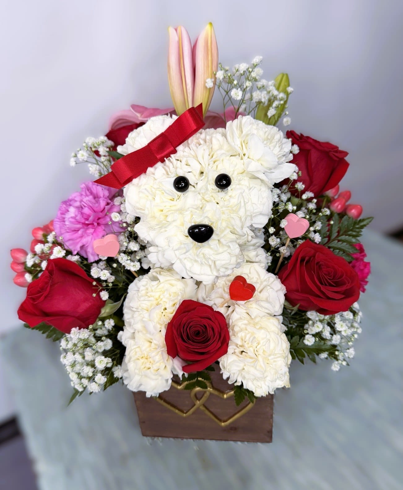 Love &amp; Romance #3 - White puppy designed out of carnations with  red roses that surround the puppy along with pink carnations, pale pink blooms, purple filler flowers and tiny white baby's breath in this Love &amp; Romance flower arrangement-perfect as a romantic bouquet or Valentine's Day gift. The tall rose focal points give the design a classic, heartfelt feel while the layered mix of pinks, purples and whites keeps the mood cheerful and tender, the kind of gift that makes the person you're thinking of feel truly adored.  Ideal for anniversaries, Valentine's Day, or any moment you want to say "I love you," this design suits someone who enjoys romantic, tactile gestures-whether they love classic roses or a sweet, smiling floral keepsake. Size and care mirror standard mixed-flower arrangements: keep blooms in fresh area and avoid direct heat to extend the life of the flowers.