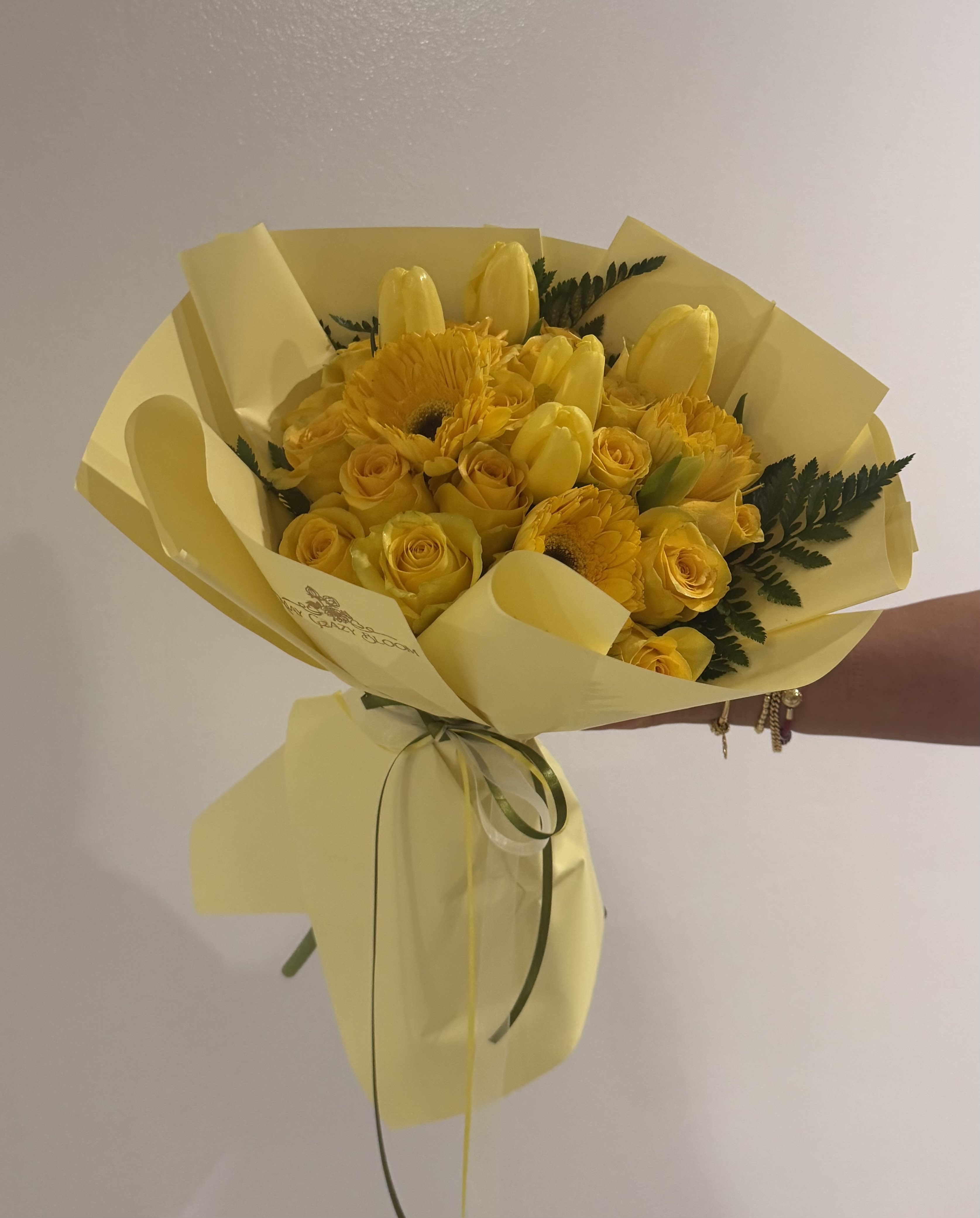 Renew  - Sunny yellow roses, tulips, and gerbera daisies come together in a compact hand-tied bouquet that reads like a burst of spring sunshine - perfect as a birthday bouquet, a cheerful thank-you gift, or a just-because pick-me-up. The bright monochrome palette immediately signals warmth and celebration, so the person you're thinking of will feel noticed and genuinely uplifted the moment they see it.  The arrangement is dense and rounded, built from vivid yellow roses, tulips, and daisy-like gerbera blooms with dark green, fern-like foliage peeking through for contrast and texture. Flowers are wrapped in layered pale yellow and floral wrapping paper and finished with a thin ribbon at the base for a clean, gift-ready presentation. Its compact hand-tied size makes it easy to carry and display on a small table or desk, bringing a fresh, sunny accent to any space.