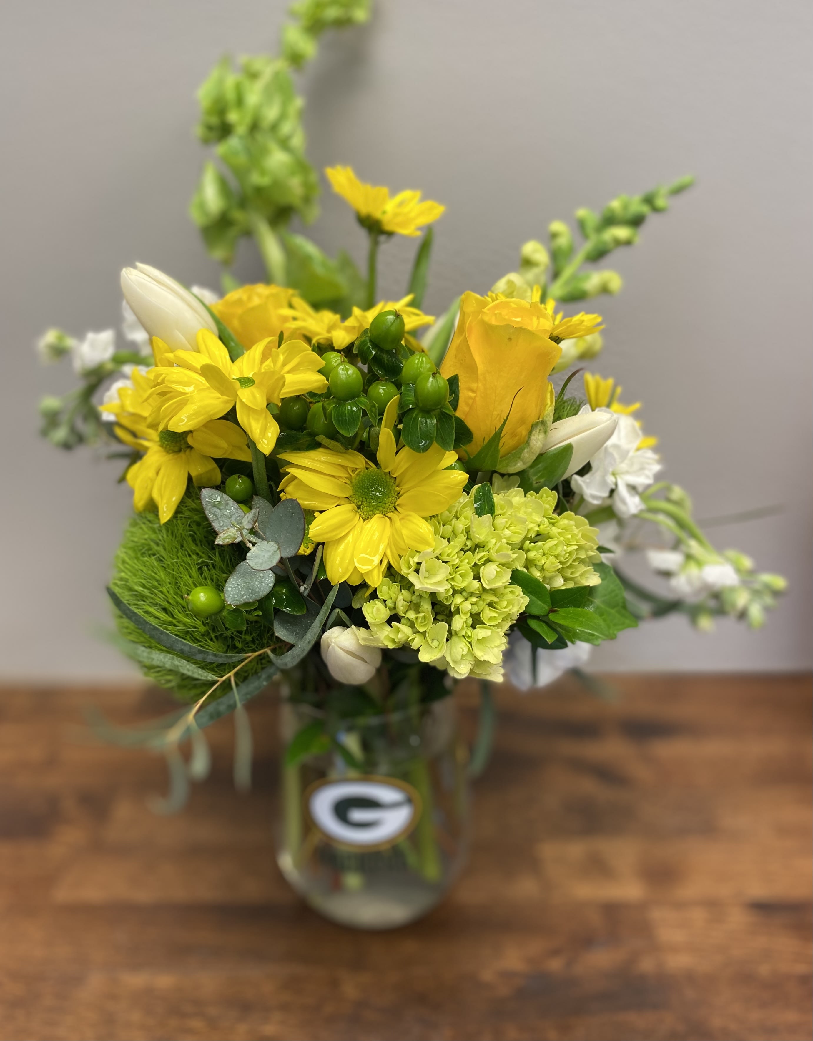 Packers for the win-e - A stemless Packers Glass wineglass, filled with a combination of green and gold (yellow) flowers with white accents.    We custom create each design based on the seasonality and availability of the flowers, so the design may look different- the value of the flowers will not be lower.  