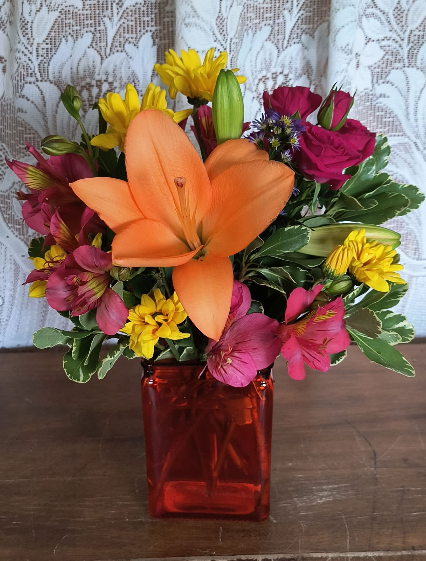 Mixed bouquet with orange lily, pink roses, yellow daisies, and a red vase