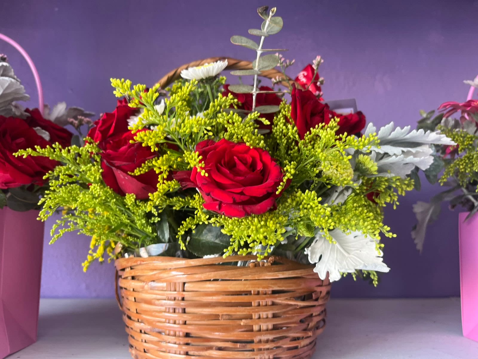 Red Rose Garden Basket - A timeless expression of love and warmth, this lush garden basket arrangement features rich crimson red roses nestled among vibrant solidago, silver-toned dusty miller, white button mums, and wispy eucalyptus sprigs — all hand-arranged in a classic woven wicker basket. The natural textures of the basket pair beautifully with the bold blooms, creating a full, cottage-garden feel that stands out in any room. Perfect for birthdays, anniversaries, Mother's Day, or simply to brighten someone's day. Available for same-day delivery throughout Chicago.