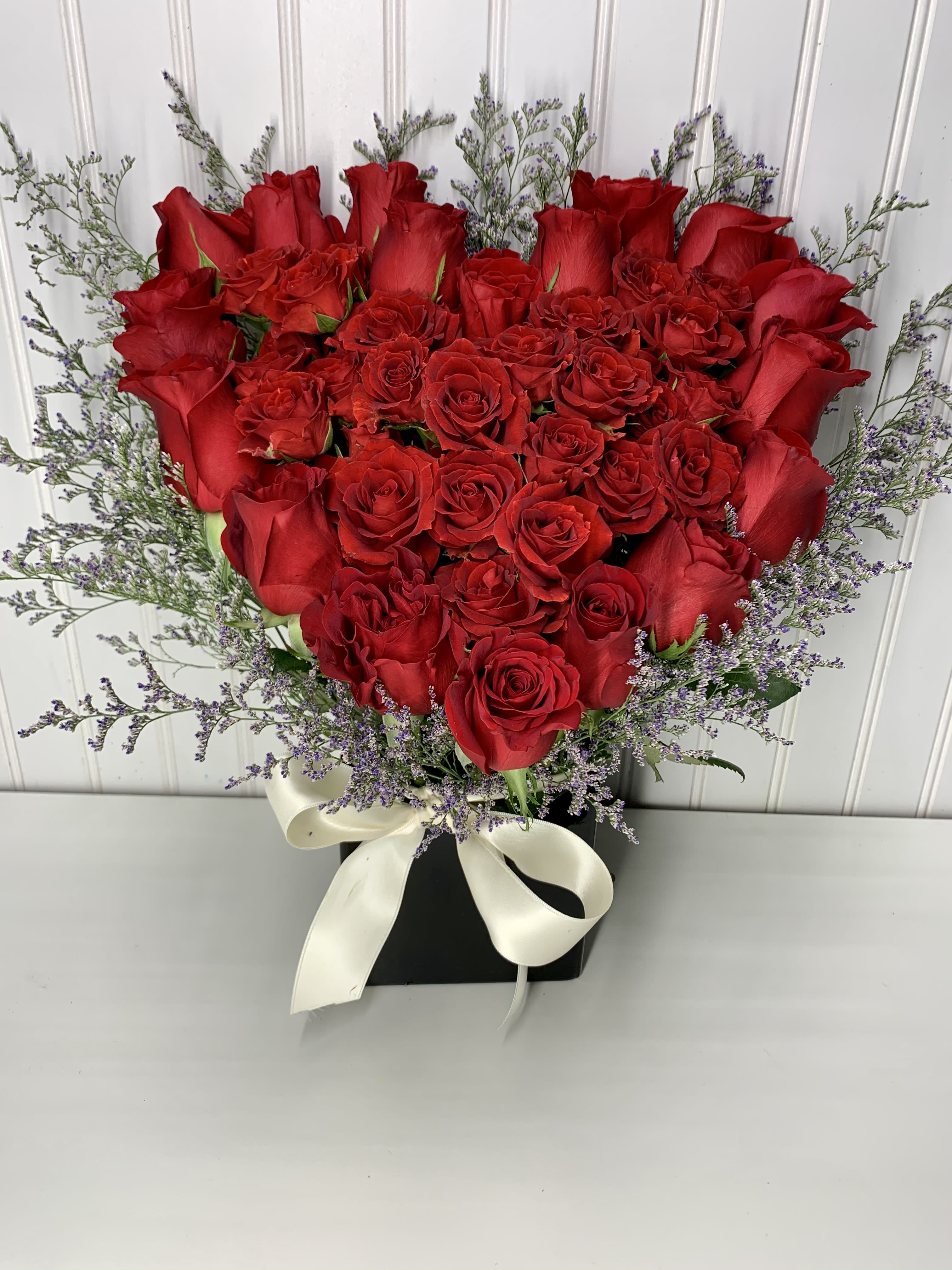 Heart-shaped arrangement of red roses in a black box with purple filler flowers and a white bow.
