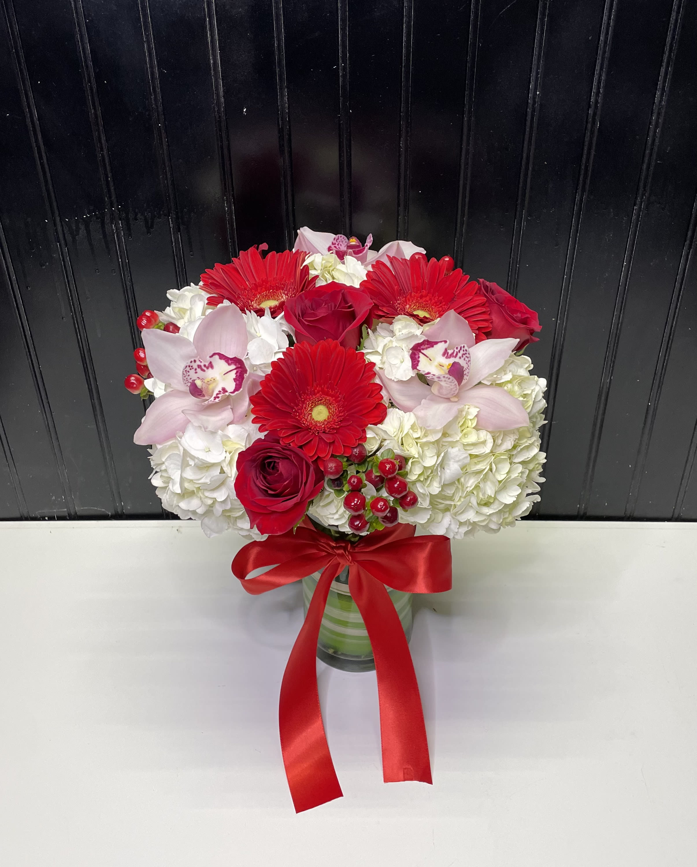 Red gerbera daisies, roses, pink orchids and white hydrangeas in a glass vase with red ribbon