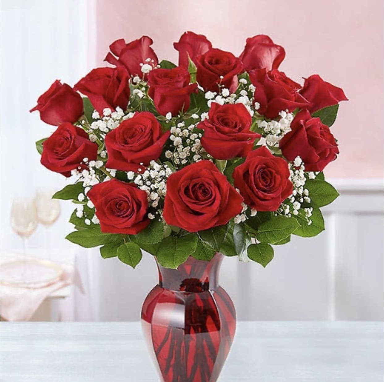 Bursting love - A lush cluster of deep red roses creates an immediate, romantic impact - the perfect anniversary or Valentine's Day flower arrangement to make her feel truly cherished. This "bursting" bouquet reads as classic and elegant, ideal for love and romance, holiday celebrations, or a thoughtful just-because surprise that says, simply, you matter.  Eighteen rich red roses are tightly grouped for a full, rounded silhouette, with small white filler blooms woven throughout to add delicate contrast and soft texture against the velvety petals. The stems are presented in a curved translucent red glass vase that mirrors the rose color for a cohesive, polished look that works beautifully for Women's Day or any special moment.  Give this to the person you're thinking of when you want them to feel celebrated and seen - whether marking an anniversary or surprising them on an ordinary Tuesday. Vase-arranged for immediate display; trim and fresh water will extend enjoyment.