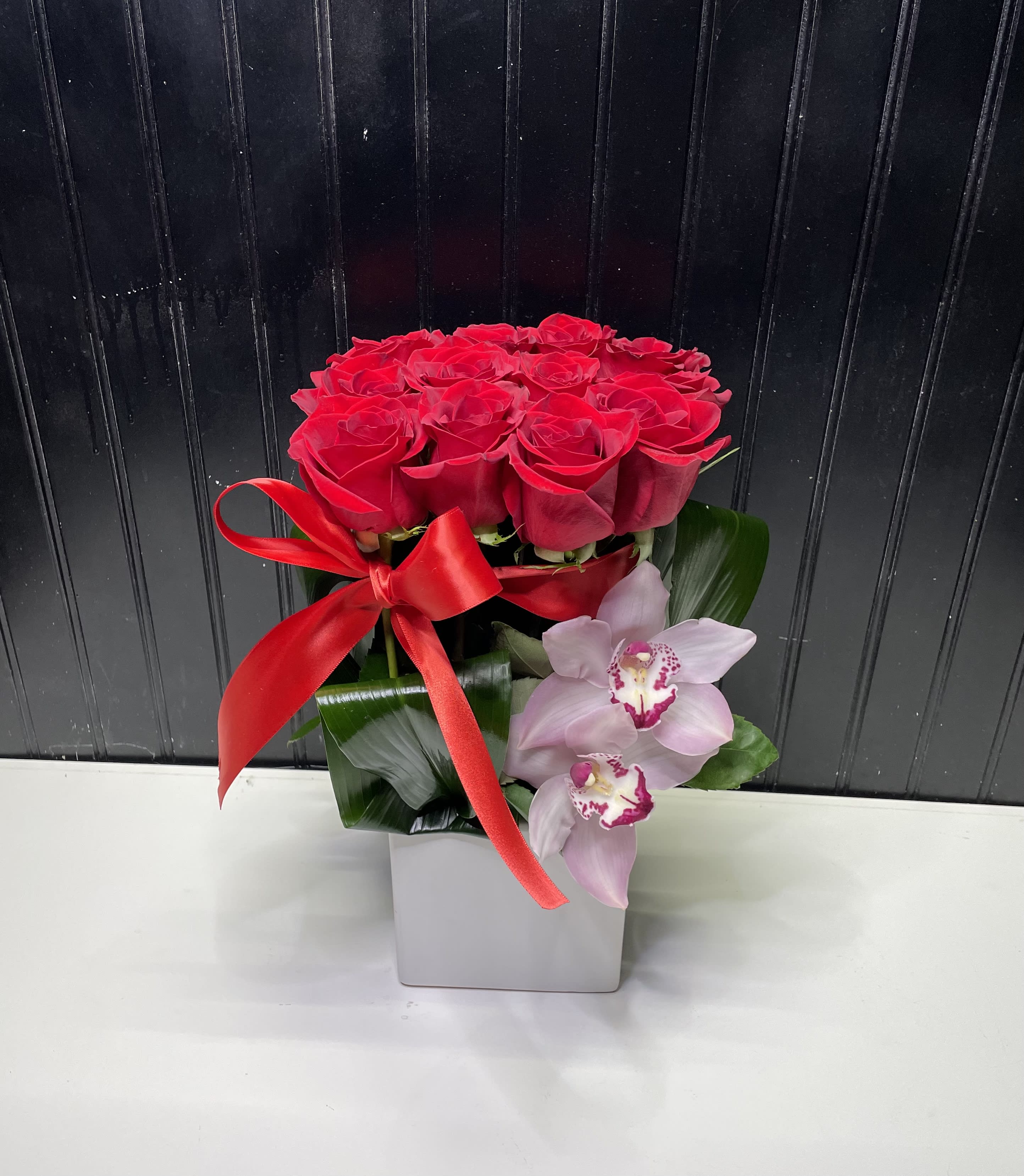 Compact arrangement of red roses with pink orchids in a square white vase with a red ribbon bow
