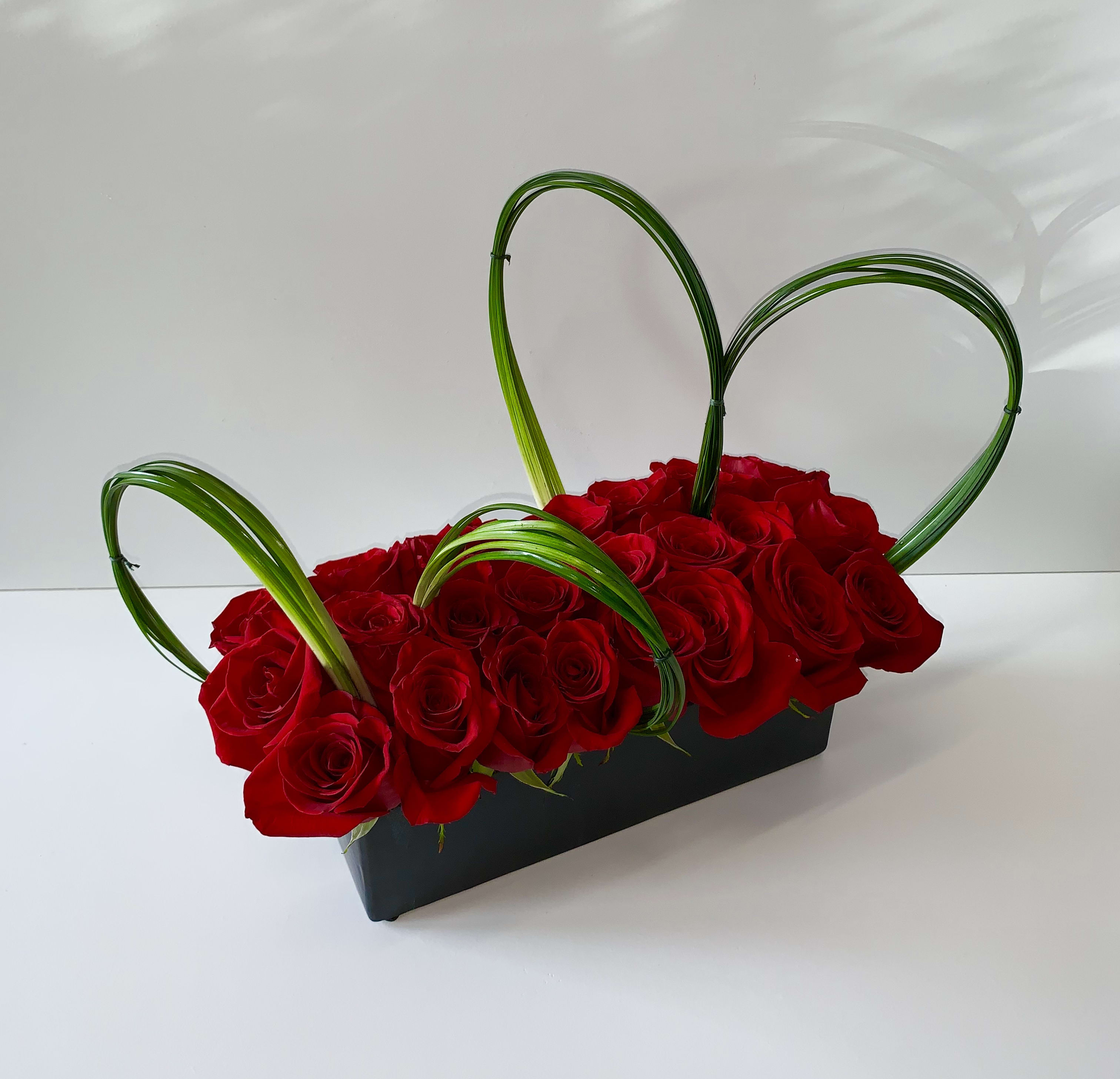 Rectangular arrangement of red roses in a black container with looped green blades forming hearts.