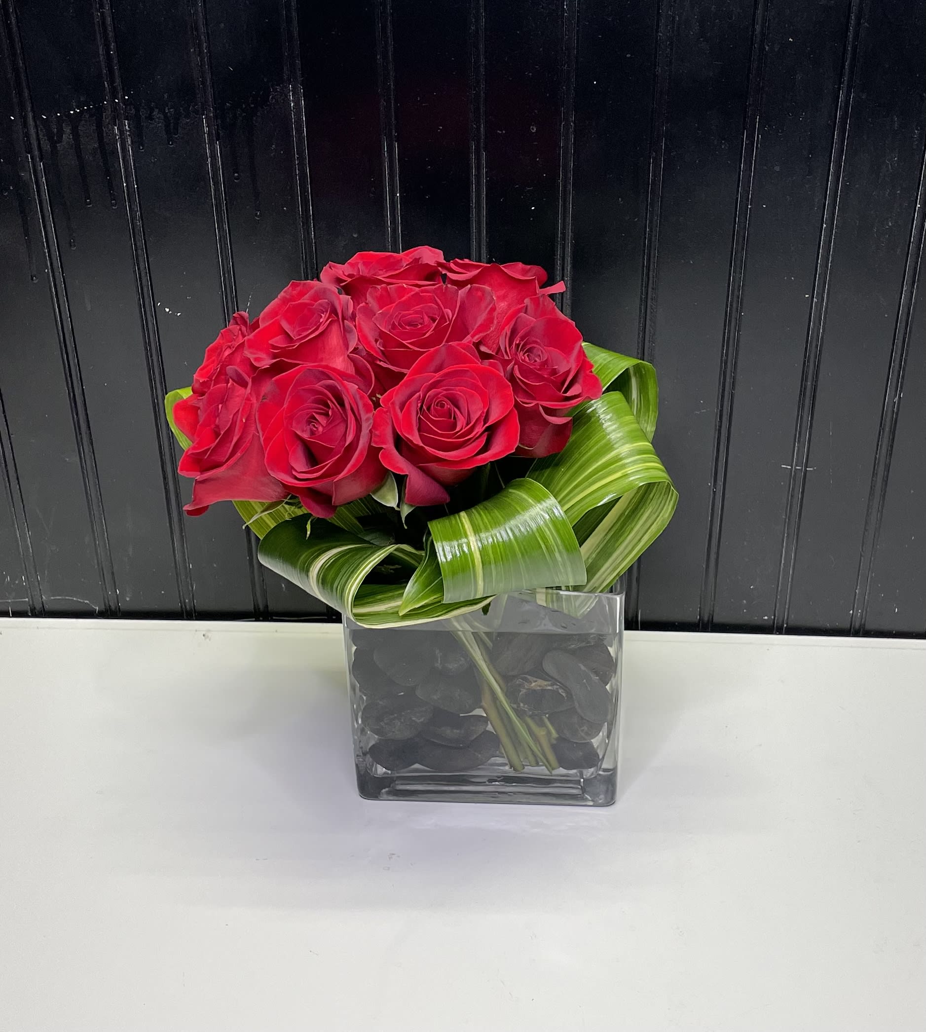 Compact arrangement of red roses with looped green foliage in a square glass vase with black stones