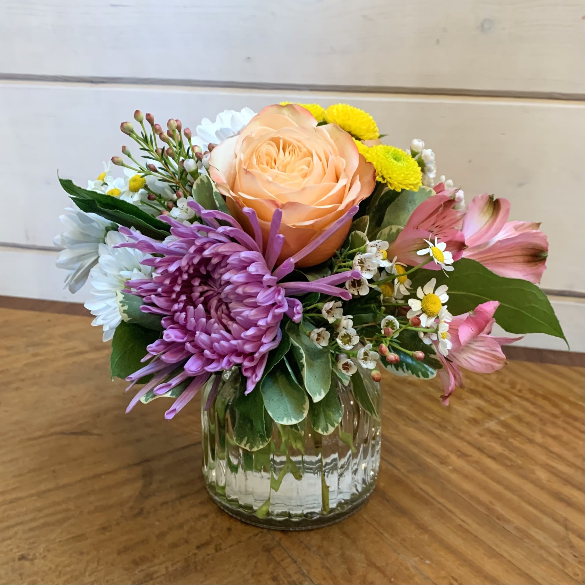 Petite Blooms - Apricot roses and a large lavender chrysanthemum with white daisies, yellow button chrysanthemums and pale pink alstroemeria in a petite medium ginger vase with seasonal filler flower. Approximately 6" by 6".
