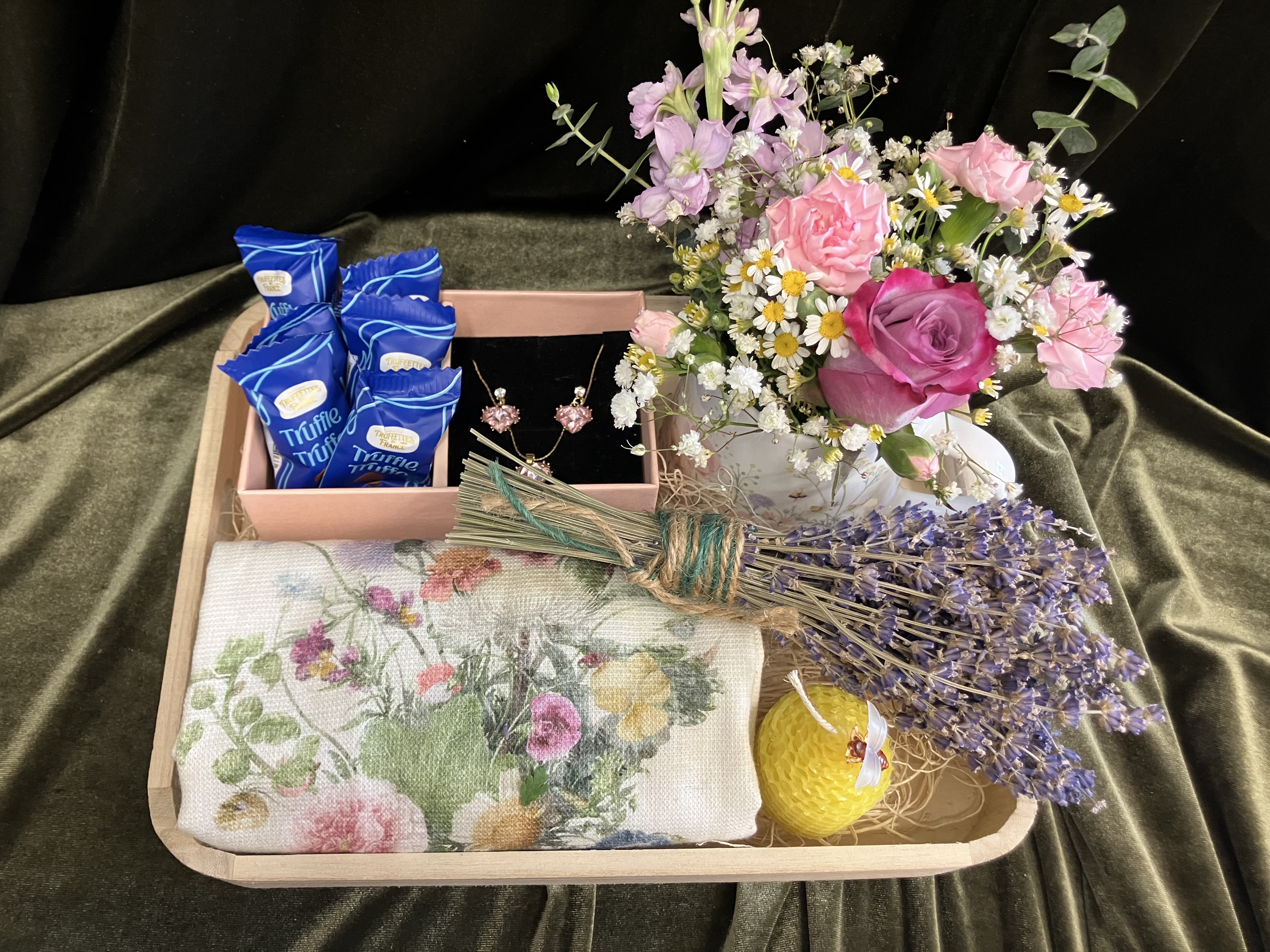 Summer Flowers and Gifts  - This flower arrangement with treats is designed to make the recipient feel genuinely celebrated: the playful mix of carnations, daisies, and other seasonal blooms creates an uplifting, fresh-scented display that reads as both joyful and thoughtful for get well, housewarming, retirement, or thank-you moments.  Arranged on a shallow wooden tray, the blooms are gathered in a pretty floral mug and paired with a marching kitchen towel, a curated set of keepsakes include chocolate truffles, a small beeswax candle, dried lavender and a pink gem necklace and earring set. The tray makes it easy to present or place on a coffee table; bouquet size and flower mix are arranged to look abundant without overwhelming a small space.  This is the kind of all-in-one gift set that works for spring celebrations or a spontaneous "just because" surprise: she'll feel seen when she opens it. Water daily for the fresh stems will keep blooms looking their best for days.