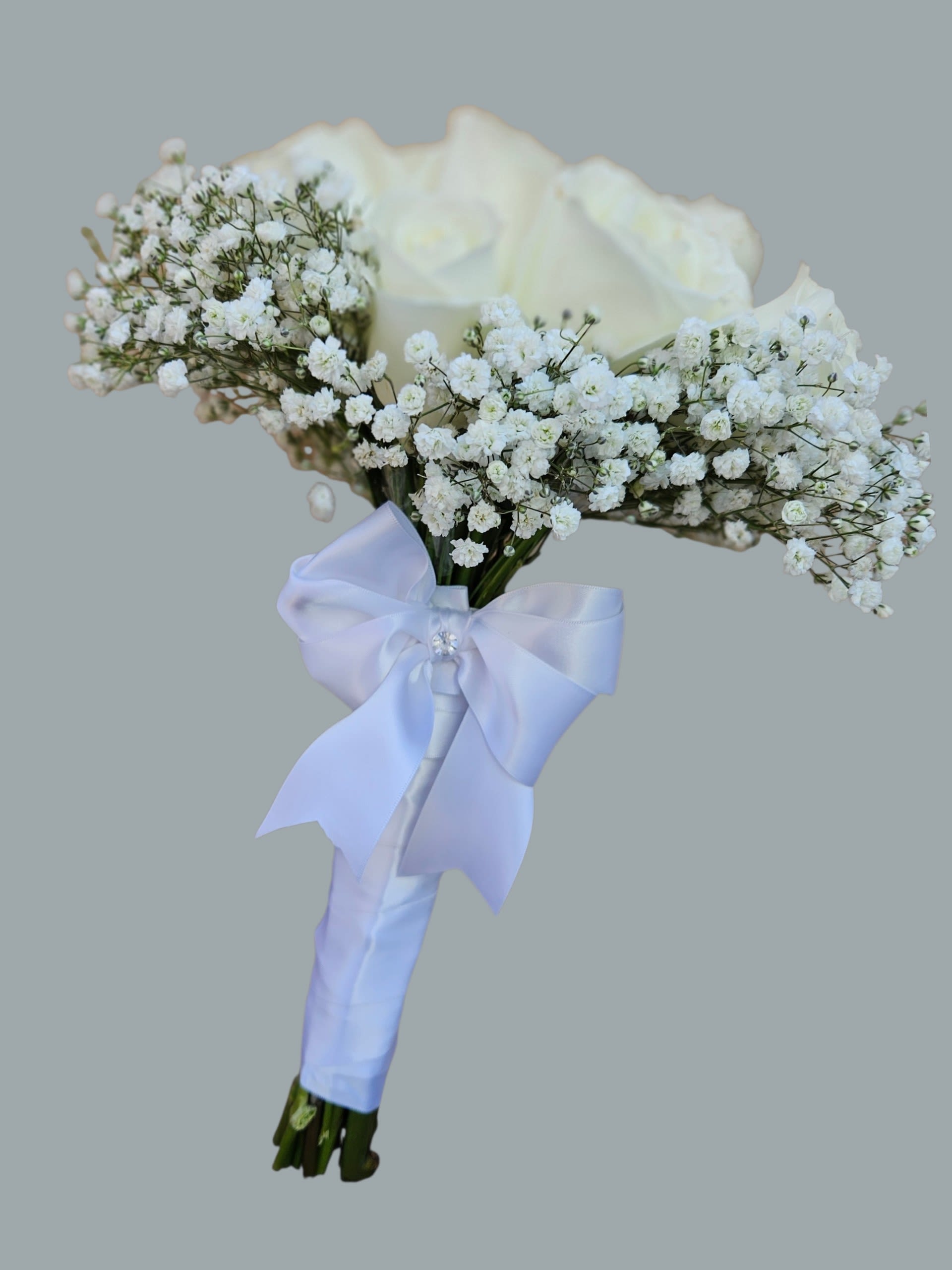 Novia Blanca  - A single large white rose crowned by clouds of baby's breath makes a quietly unforgettable bridal bouquet - perfect as a wedding bouquet or for any ceremony where she should feel luminous and seen. The rounded, hand-held shape and pale lavender satin ribbon give Novia Blanca a soft, formal look that reads classic and romantic; a small decorative gem pin adds just the right touch of sparkle for a bride or attendant.  This arrangement combines creamy white roses with airy clusters of gypsophila for a lace-like texture around the focal blooms. Stems are wrapped in pale lavender satin ribbon and finished with a jeweled accent; colors include white, lavender, and touches of green from the foliage for a fresh, elegant palette. The size is a compact, rounded hand-tied bouquet meant to be carried comfortably down the aisle.  Made to flatter photos and feel effortless in the hand, Novia Blanca is designed for brides, bridesmaids, or any ceremony where purity and romance matter. Order with confidence for weddings or formal events - simple care includes keeping stems in water until the moment of carrying and storing in a cool place before use.