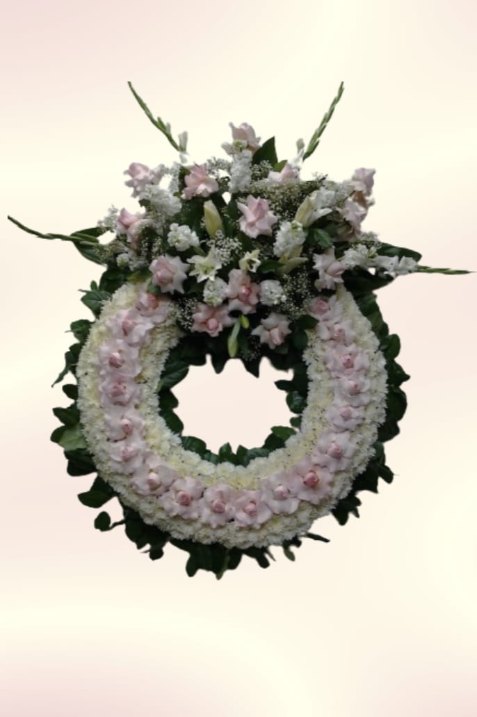 Soft Pink &amp; White Wreath - A soft halo of pale pink and white creates a gentle, reverent presence with this floral wreath-perfect for sympathy or memorial services. The circular arrangement reads as a comforting tribute, the kind that quietly honors a life and lets the person you're remembering rest at the center of the moment.  Dense white blooms form the base of the large, hollow-centered wreath while pale pink roses, lilies, carnations and mixed blossoms gather more heavily at the top, where taller stems and varied textures rise above the ring. Dark green foliage frames the design, providing contrast to the pastel palette and lending a formal, ceremonial silhouette. The overall construction is substantial and layered, designed to maintain its shape when displayed on an easel or laid flat.  This wreath offers a respectful way to send condolences and support-appropriate for funeral services, memorials, or as a lasting tribute at a graveside. For peace of mind, please confirm delivery details and placement instructions when ordering so the arrangement arrives ready to present.
