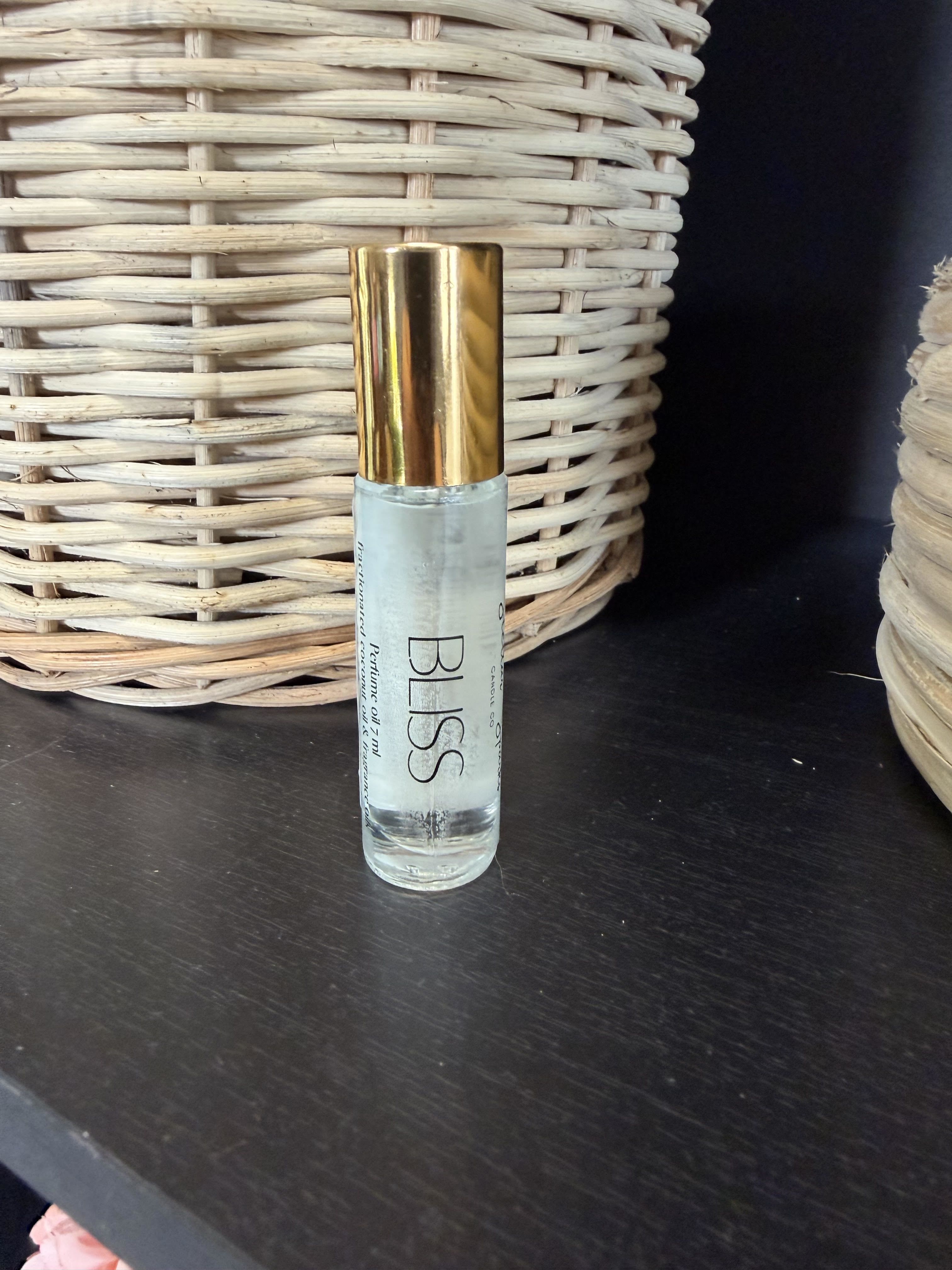 Bliss Perfume - A compact perfume roller perfect as a small gift or add-on for birthdays and "just because" moments, this perfume bottle reads as clean, minimalist, and thoughtfully styled. The product type is a gift-size perfume roller ideal for pairing with a bouquet for anniversaries, thank-you gestures, or a simple pampering treat for the person you're thinking of - she'll feel noticed by a personal, wearable luxury that fits in a purse.  The clear glass roller bottle features a metallic gold cap and a simple "BLISS" label, giving it a modern, understated look that complements both black and natural decor. Photographed against a dark shelf with woven wicker in the background, the presentation highlights the bottle's small, giftable scale and homey texture; it's an easy add-on to a floral arrangement or a standalone token of care.