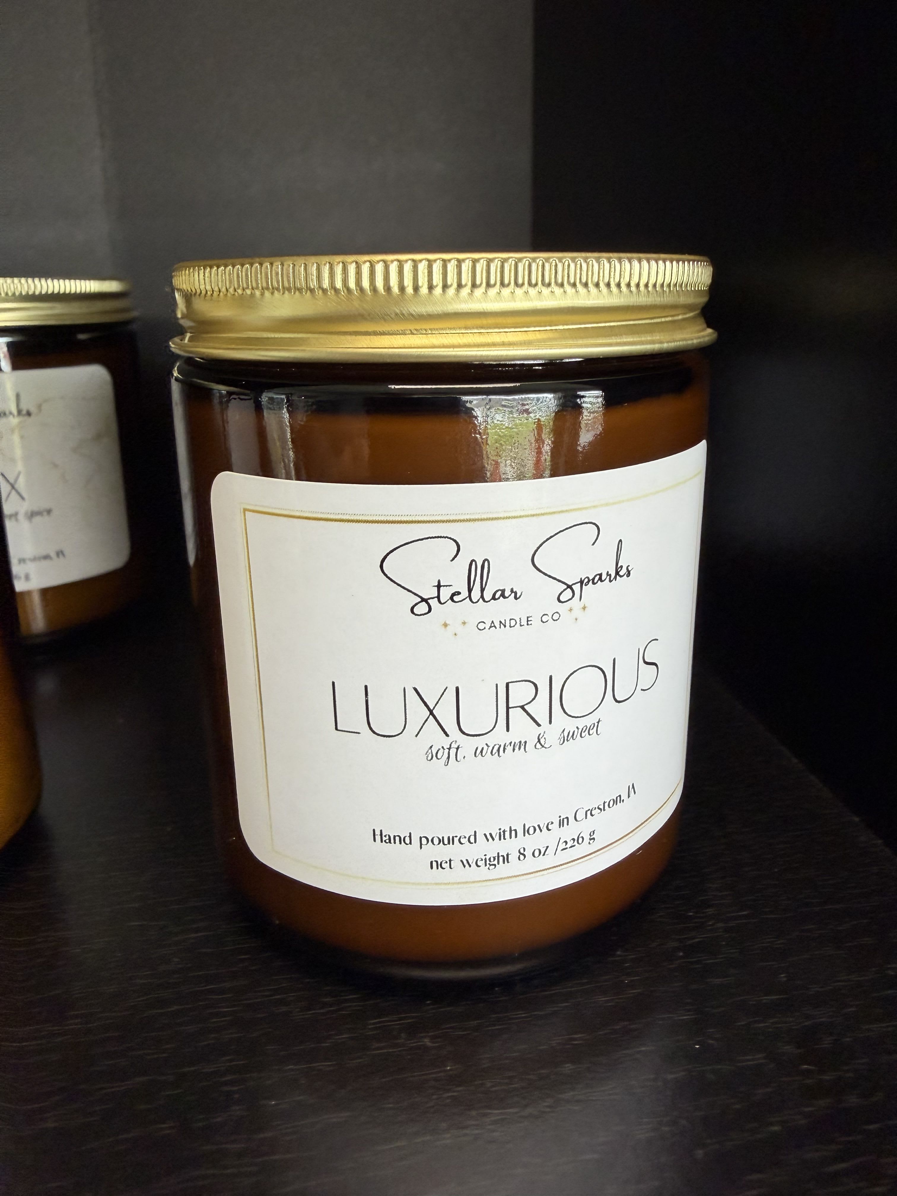 Luxurious 8oz Candle - Warm, cozy fragrance meets polished design in this candle arrangement-perfect as a thoughtful gift or a stylish addition to a sympathy or housewarming moment. The 8oz glass jar candle arrives in a rich amber vessel with a gold metal lid and a clean white label that reads "LUXURIOUS," signaling a soft, warm, sweet scent meant to soothe and comfort the person you're thinking of.  The candle's palette-amber glass, white label, and metallic gold accents-creates a neutral, elegant look that fits modern and classic interiors alike. Housed in a sturdy glass jar with a snug metal lid and clear product labeling, it's an easy, tasteful choice when you want to send calm and warmth without fuss.  Ideal for sending to someone settling into a new home, marking an anniversary with understated luxury, or offering gentle comfort during a difficult time, this candle arrives ready to use and enjoy. Burn on a stable surface, trim the wick to 1/4" before each lighting for an even burn, and keep away from drafts and flammable items for best results.