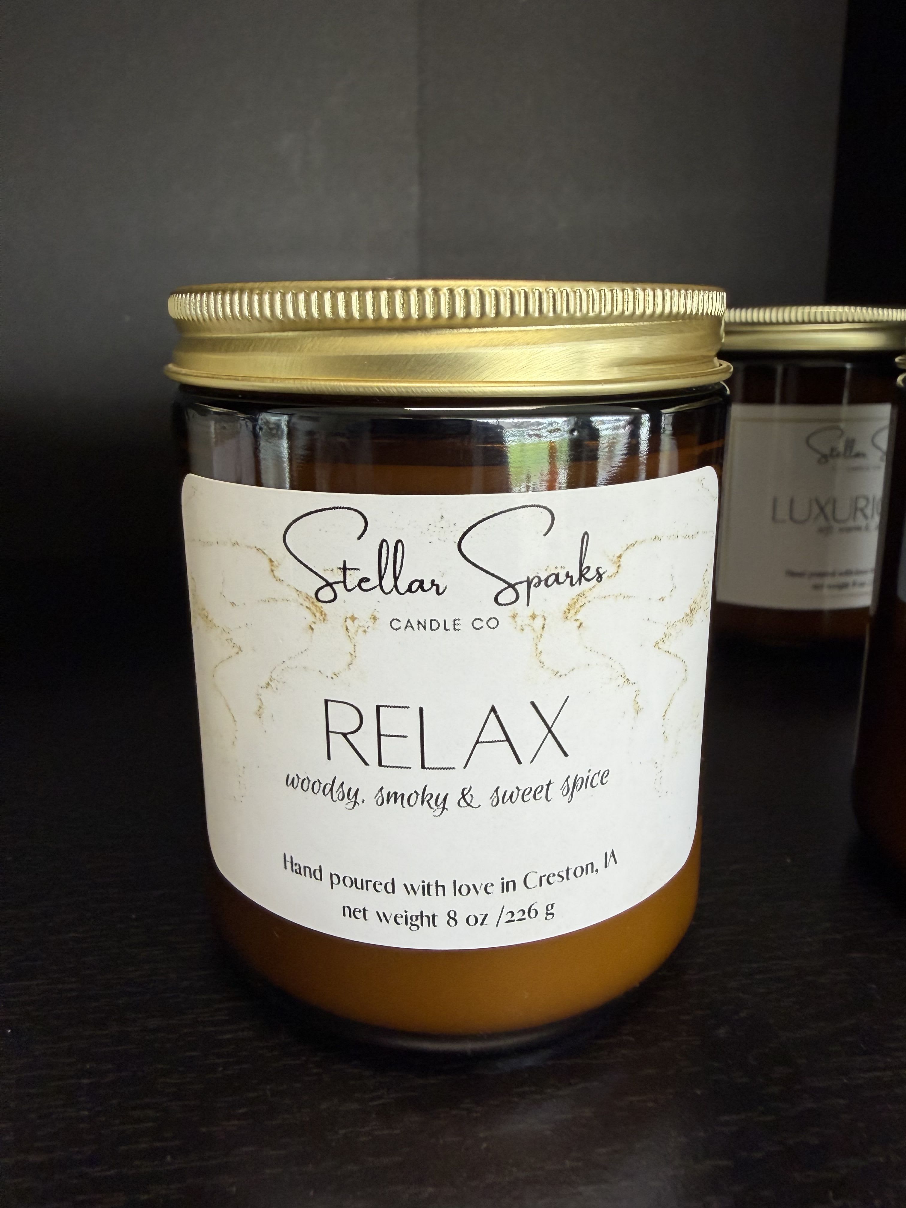 Relax 8oz Candle - Warm amber glass and a calm, cozy scent make this Relax 8oz Candle an ideal gift for home moments and self-care rituals. As a home fragrance gift or part of a curated spa set, this scented candle communicates "relax" at a glance-perfect for birthdays, housewarmings, or as a thoughtful way to say you're thinking of someone who needs a peaceful pause.  The candle comes in an 8oz amber glass jar with a gold metal lid and a white front label printed in bold black type that reads RELAX, with smaller copy noting a woody, smoky and sweet spice fragrance profile. The clean, minimalist presentation pairs effortlessly with white, black, or natural décor; a second jar shown nearby suggests it fits well in a collection or paired gift set.  Give it to someone who could use a moment of calm-the smoky-woody notes warm a living room or bedside table without overpowering. Standard candle care applies: trim the wick before each burn and allow a full melt pool on first use to prevent tunneling.