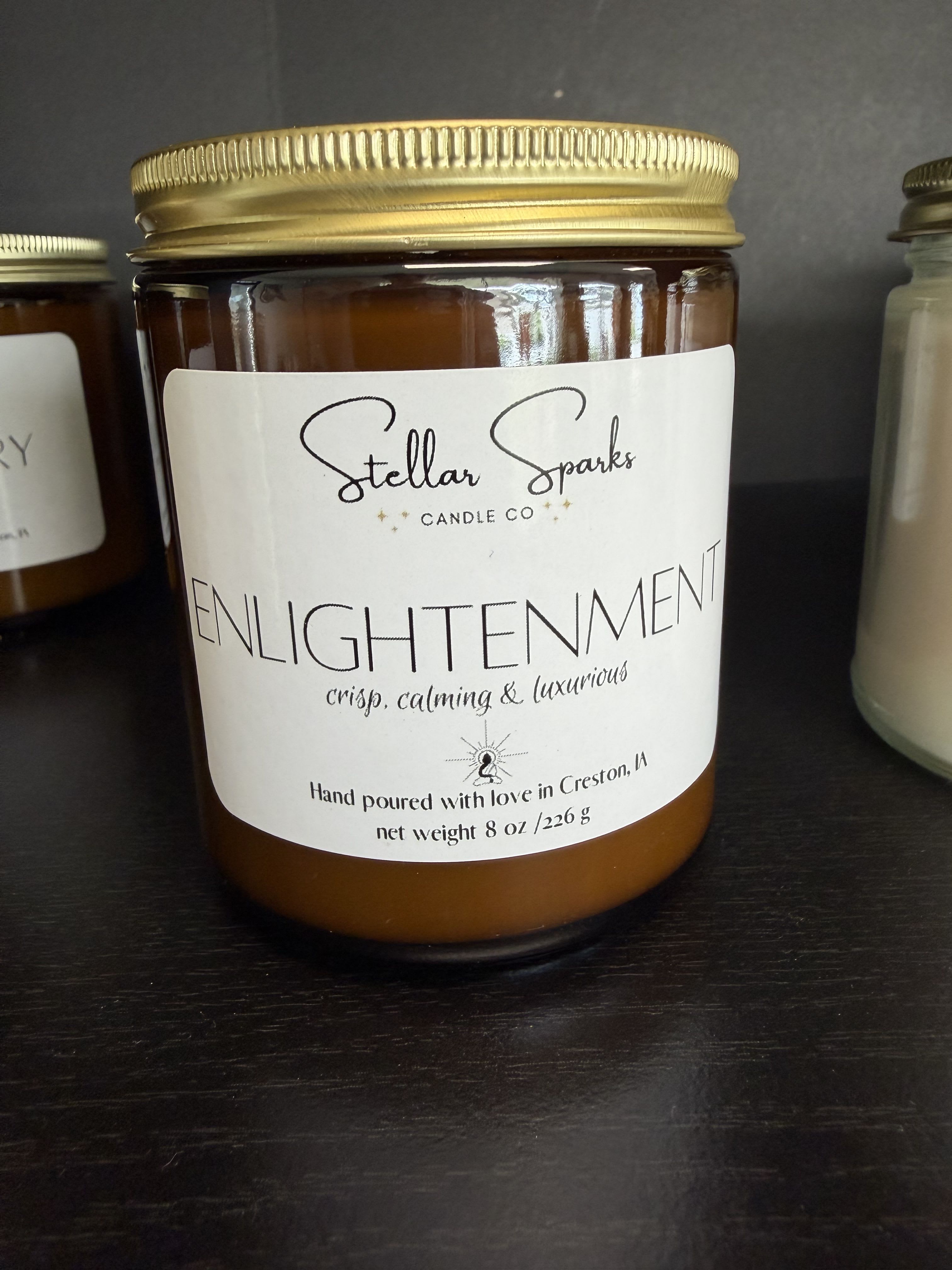 Enlightenment 8oz Candle - An elegant amber glass candle for everyday gifting and self-care, the Enlightenment 8oz Candle makes a quiet, modern statement-perfect as a housewarming gift or a thoughtful accompaniment to a birthday, thank-you, or "thinking of you" moment. The soothing, minimalist look reads calm and deliberate; the person you're thinking of will feel seen by a stylish, useful present that adds warmth without overpowering a room.  The candle comes in a compact 8oz amber glass jar with a gold metal lid and a clean white label from Stellar Sparks Candle Co., giving it a neutral black, white, and warm-amber palette that fits many decor styles. Sized to sit on a bedside table or bathroom shelf, it's packaged simply for immediate display and everyday use.