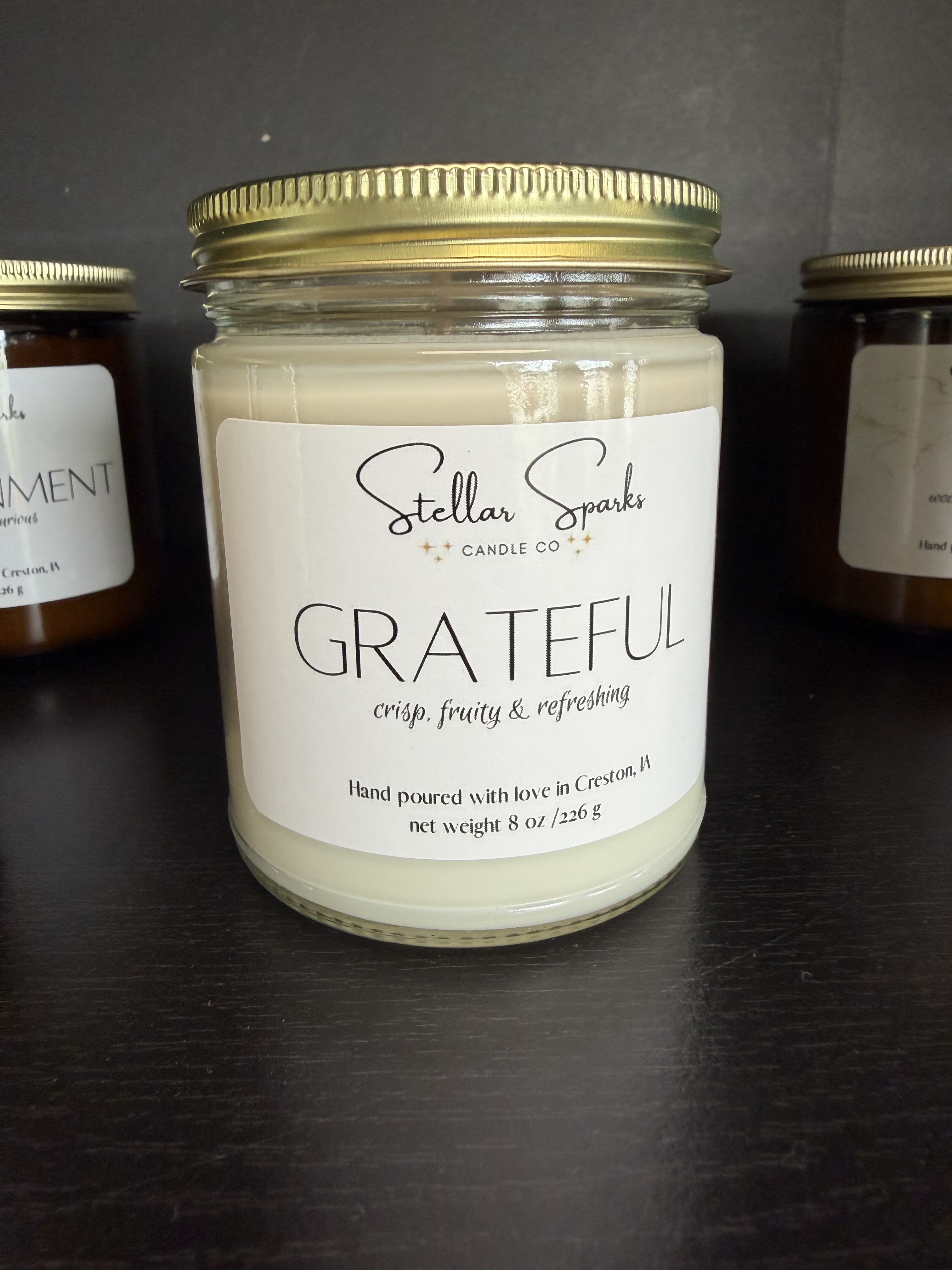 Grateful 8oz Candle - A serene, gift-ready jar candle that makes a thoughtful accompaniment for birthdays, thank-you gifts, or a cozy housewarming moment. This 8oz glass jar candle labeled "GRATEFUL" sets a reflective, calming tone-perfect for the person you're honoring when you want them to pause and feel appreciated.  The clear glass jar holds pale cream wax topped with a gold metal lid and a simple white label that reads GRATEFUL with subtle branding and a scent note beneath. Clean, handmade styling and neutral white, black, and gold accents let this candle slip easily into any decor-on a bedside table, bathroom shelf, or coffee table vignette.  Give it as a small but meaningful gesture to say thanks, congratulations, or thinking-of-you; it's the kind of present that invites quiet moments. 8oz size makes it easy to ship or tuck into a gift box; burn on a heat-safe surface and trim the wick to 1/4" before lighting for best results.