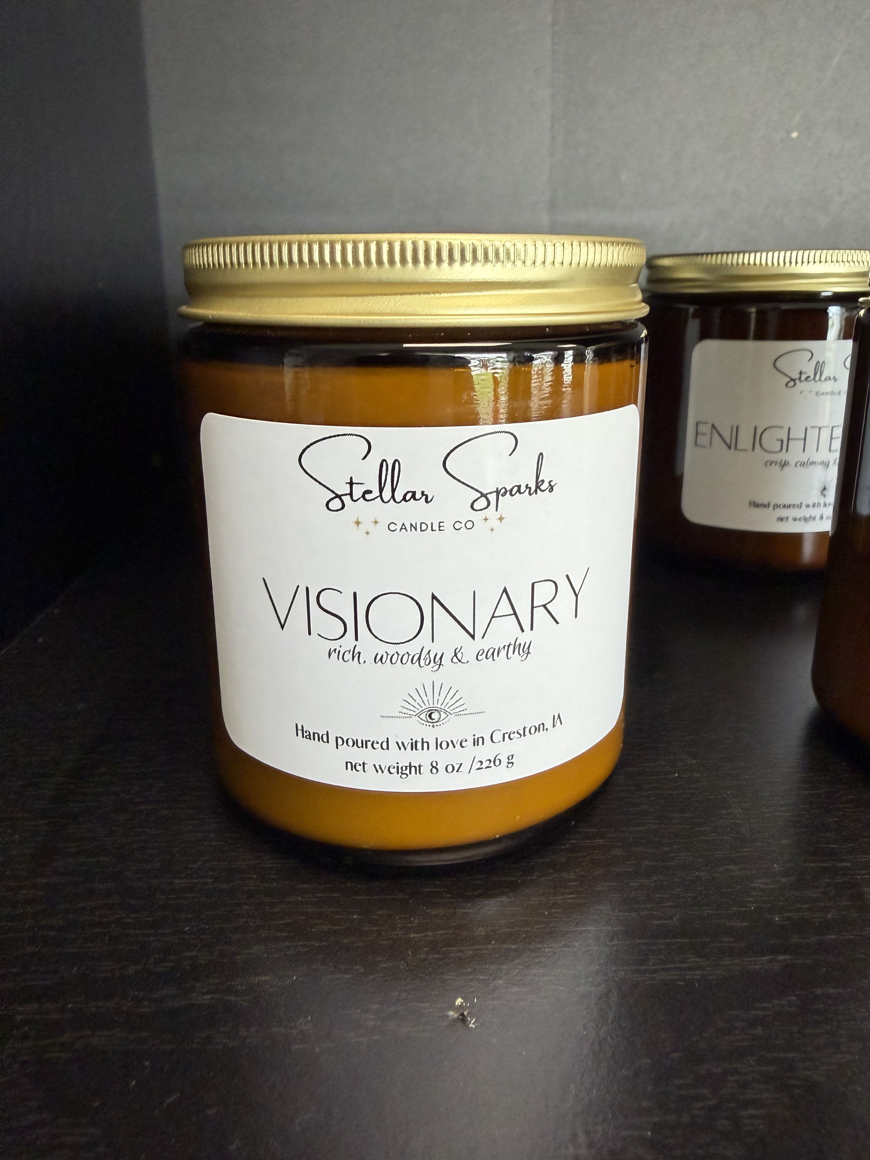 Visionary 8oz Candle - Warm amber glass and a rich, woodsy scent make this 8oz candle a thoughtful gift for housewarmings or to mark a cozy anniversary evening. The scented candle in an amber glass jar with a gold metal lid and clean white "Visionary" label reads "rich, woodsy &amp; earthy," signaling a grounded, grown-up fragrance that will make the person you're thinking of feel settled and cared for.  The candle's palette of orange, white, black, and yellow is expressed through the amber jar, the white label, and the gold lid, creating an artisanal, minimalist presentation that works well on mantels, bedside tables, or paired with flowers. Housed in a sturdy glass jar with a metal lid and tasteful label, the 8oz size is ideal for moderate burn time without taking over a room-perfect for gifting as a solo present or as part of a coordinated home-fragrance set.  Give this scent to someone who appreciates calm, earthy aromas and thoughtful design; it reads as intentional and refined rather than flashy. Order for delivery or pickup and suggest pairing with a simple bouquet for a complete, comforting gift-no special care is needed beyond the usual candle-safety practices.