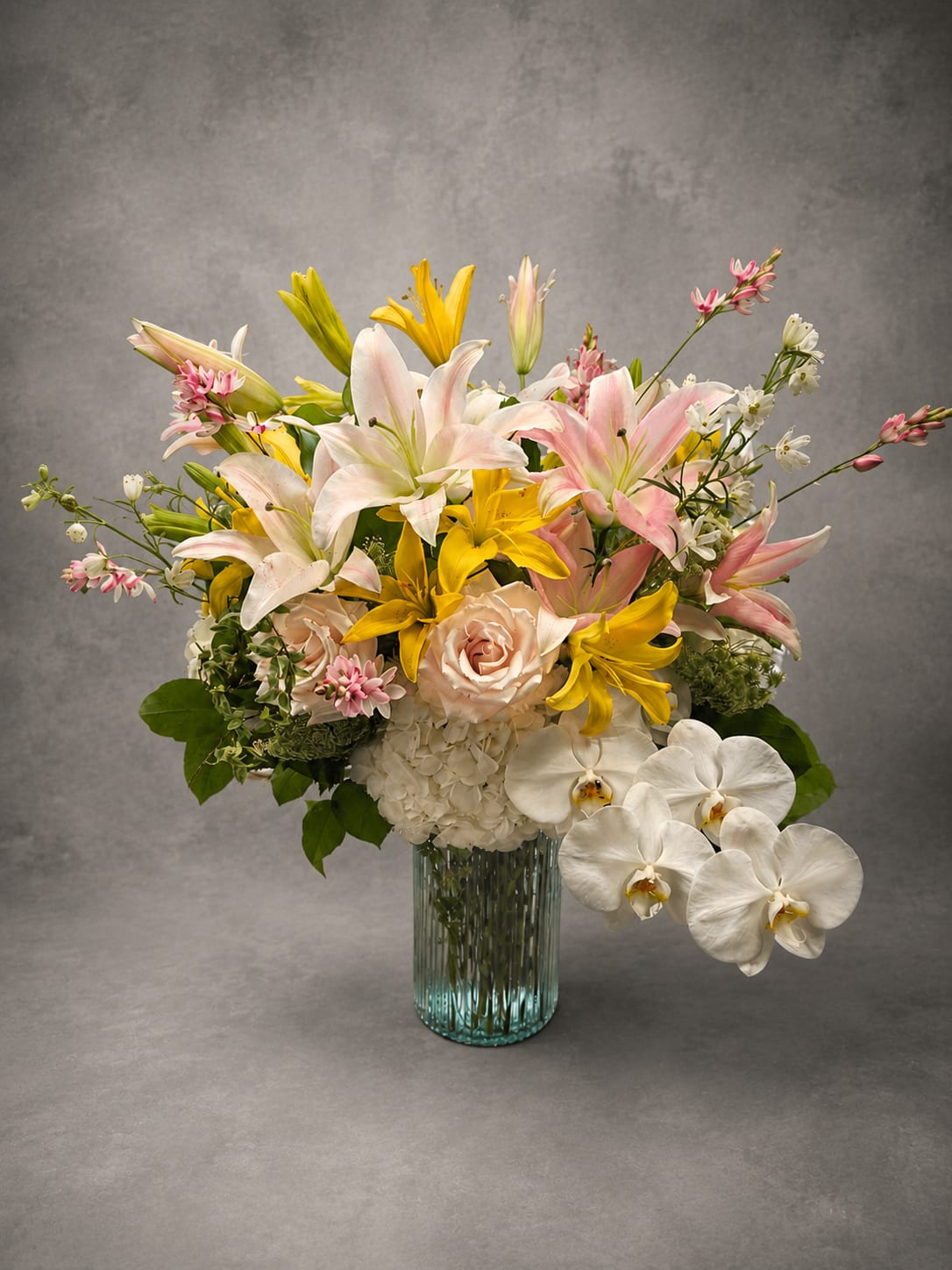 Blushing Lilies - An elegant gathering of premium lilies, blush roses, white hydrangea, and seasonal accents arranged in a modern glass vase. Soft colors and lush textures create a design that feels romantic, refined, and effortlessly beautiful.