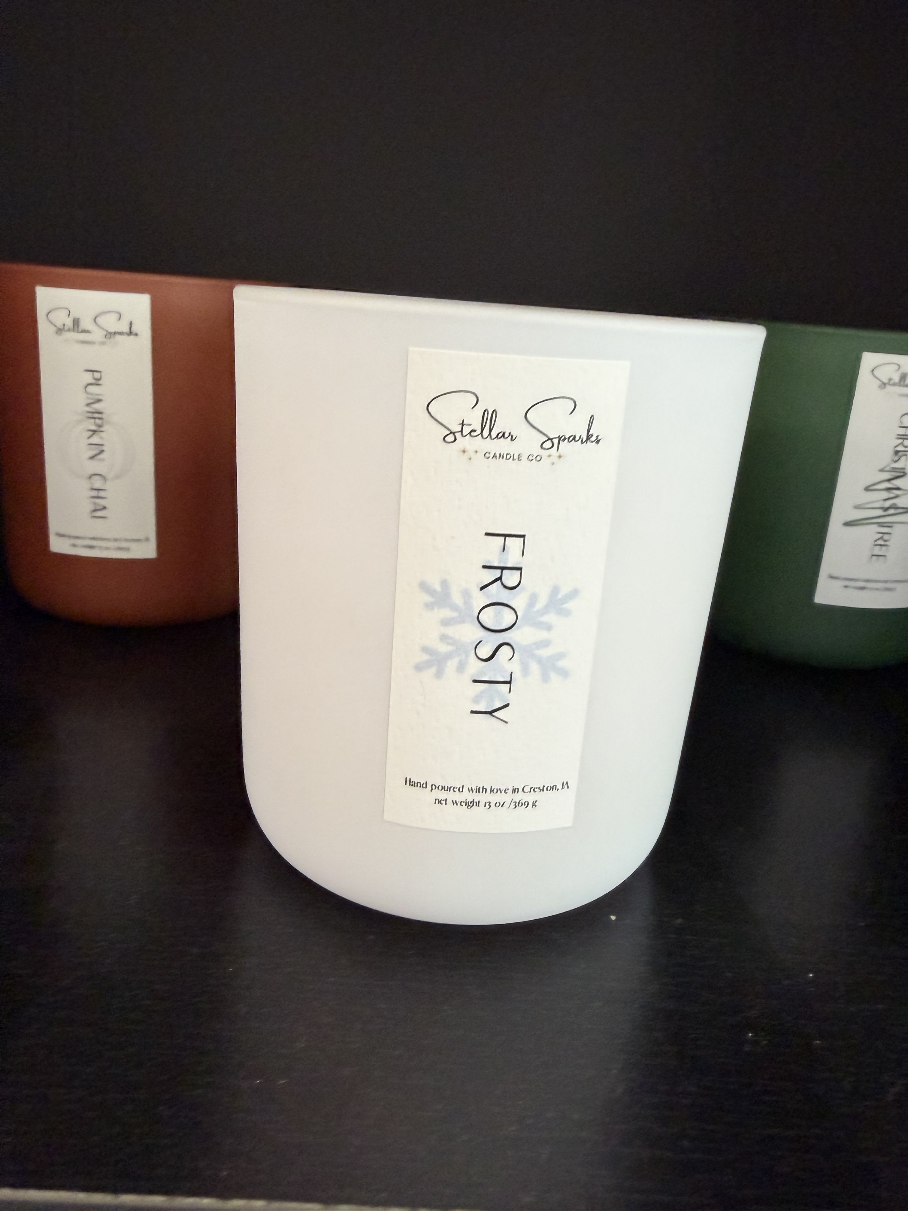 Frosty 13oz Candle - A trio of seasonal scented candles arranged for gift-giving brings a cozy, holiday-ready vibe - perfect as a holiday gift or stocking-stuffer for someone who loves festive scents. This flower arrangement page features the Frosty 12oz Candle alongside two complementary scents, positioned as a thoughtful seasonal set for Christmas or winter gatherings.  Each candle comes in a simple, clean container with clear product labels: the central Frosty 12oz white candle is flanked by a red Pumpkin Chai and a green Christmas Tree scent. The white, red, and green color trio reads classic and cheerful; containers are retail-friendly and ready to display on a mantel or dining table. Though presented with minimal floral accents, the set pairs beautifully with winter foliage if you'd like to create a quick centerpiece.  Give this to someone who appreciates warm, familiar aromas - the kind of gift that makes a room feel welcoming and celebrated. Candles arrive labeled and ready to use; for best results burn on a heat-safe surface and follow standard candle-safety practices.