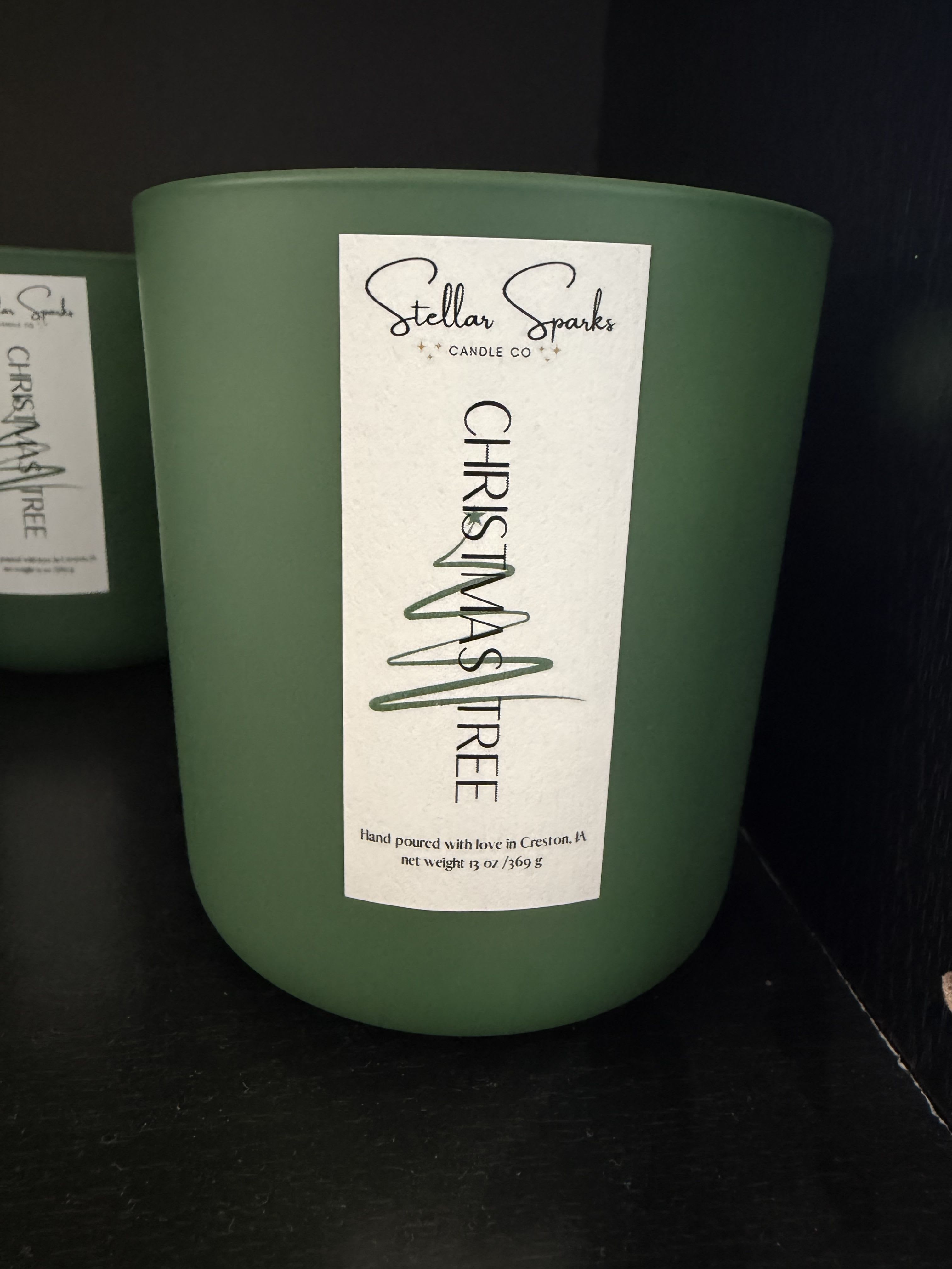 Christmas Tree 13oz Candle - A deep forest-green candle that reads like a cozy winter evening-this Christmas Tree 13oz candle is a seasonal gift and table-ready holiday accent. Perfect as a holiday gift or for seasonal home decor, the matte green cylindrical container and tall white label give a modern, minimalist look that makes the recipient feel warmly remembered and festive.  The 13oz candle arrives in a sleek matte green glass jar with a contrasting tall white product label marked "Stellar Sparks Candle Co" and "Christmas Tree," and a simple black product label element for a refined, moody finish. Clean lines and muted colors-green, white, and black-let it sit effortlessly on mantels, entry consoles, or as part of a holiday centerpiece without competing with other decorations.  Give this to someone who loves winter scents and understated style-the kind of gift that says you thought about their home and comfort. Sized for multiple evenings of burn, it's easy to place and enjoy; order as a single-season treat or pair with other seasonal pieces for a fuller holiday display.