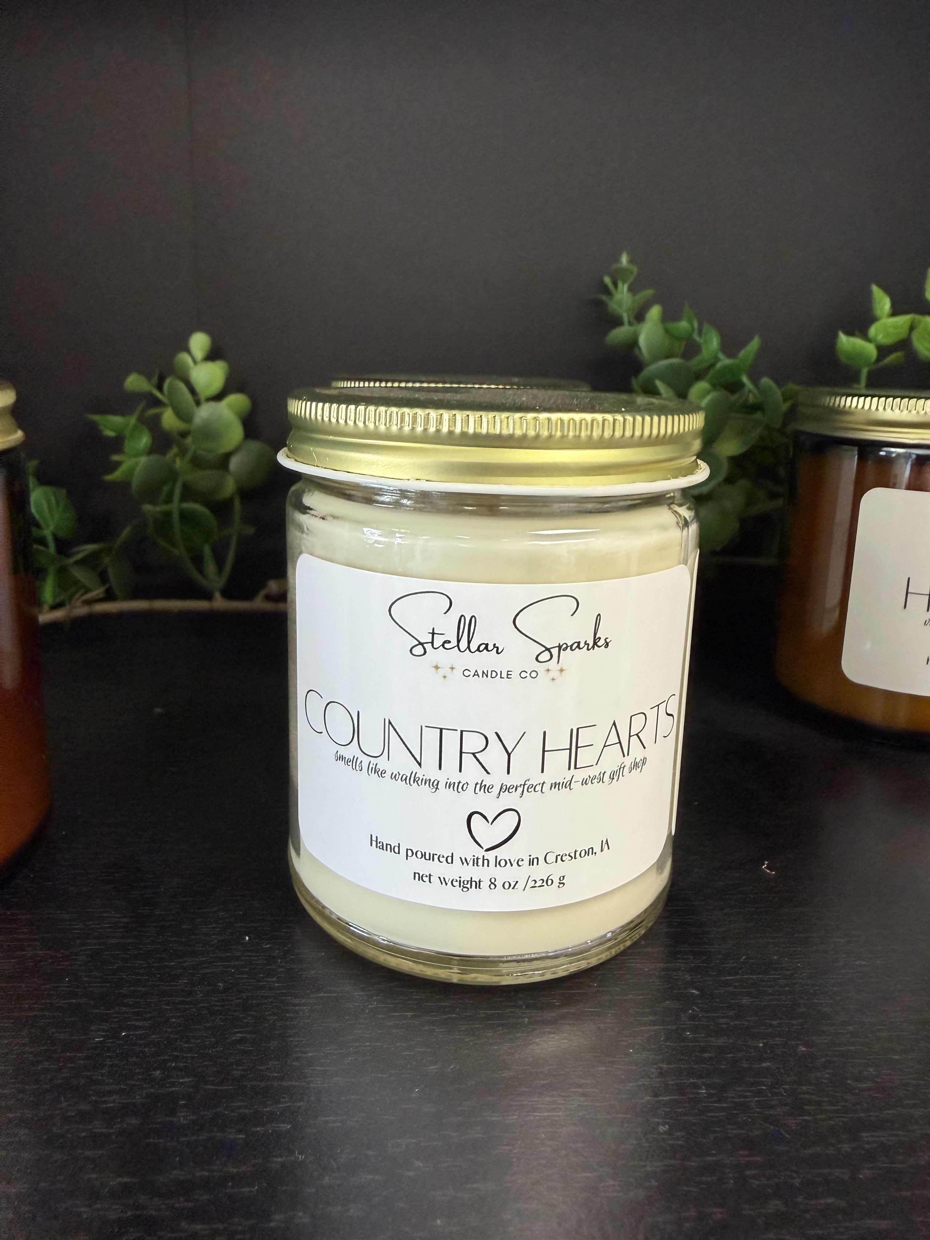 Country Hearts 8oz Candle - Pale cream wax, a gold metal lid, and a simple white label combine into a cozy, rustic gift that feels like a warm kitchen hug - the perfect candle for Mother's Day, housewarming, or thinking-of-you moments. As a flower arrangement accent or standalone present, this glass jar candle brings a calm, country-inspired note to the room and helps the person you're sending it to feel seen and comforted.  The Country Hearts 8oz candle sits in a clear glass jar topped with a gold metal lid and a white front label for a clean, timeless look. Soft cream wax paired with small green sprigs arranged behind the jar creates an understated natural palette of white, yellow, black, and green accents that complements farmhouse or cottage decor. Sized for personal use, it's an easy add-on to a bouquet or a thoughtful gift on its own - simple to display, easy to enjoy, and perfectly suited to bring quiet warmth to someone's everyday space.