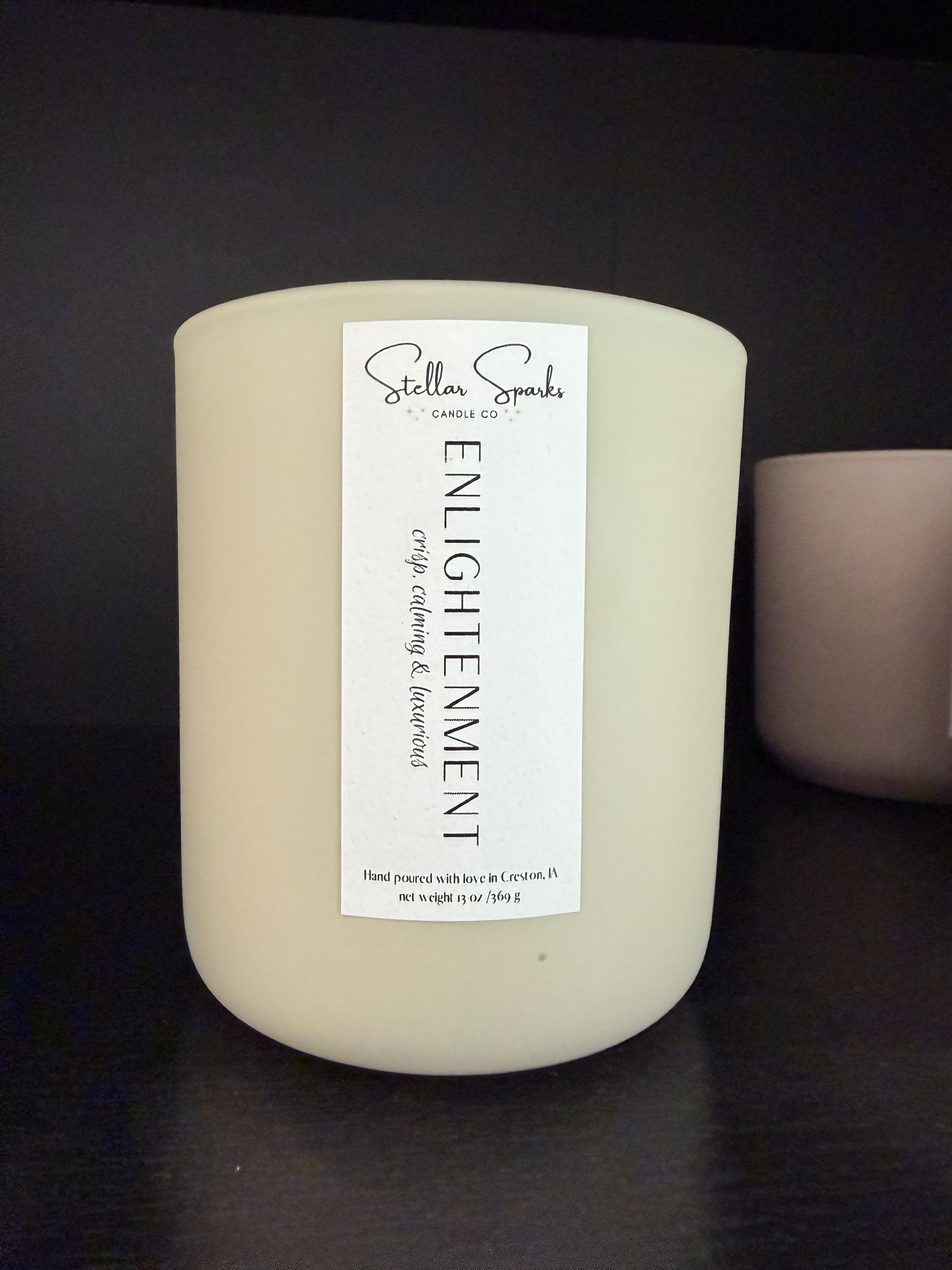 Enlightenment 13oz Candle - A refined, calming gift that reads as both home fragrance and thoughtful keepsake, this flower arrangement pairs with a 13oz Enlightenment candle for a serene sympathy or housewarming gesture. The centerpiece is the cream-colored, rounded candle jar labeled "Enlightenment"-its minimalist white label and soft matte finish create a quiet, luxurious presence that complements white and pale-pink florals for occasions like sympathy, get-well, or a gentle birthday surprise.  The arrangement features white and pale-pink blooms (and complementary greenery) chosen to echo the candle's clean, understated aesthetic; soft textures and airy shapes let the neutral candle remain the visual anchor while blooms add warmth and tenderness. The product includes the 13oz candle in its rounded jar with the Stellar Sparks Candle Co label-an elegant hard good that elevates the floral gift without overwhelming it.  Made to make the recipient feel soothed and seen, this pairing is ideal for someone needing calm or for marking a quiet milestone. Order with standard sizing; the candle offers long-burning fragrance to extend the moment, and simple care for the flowers-fresh water and a cool spot-keeps the gift looking its best.