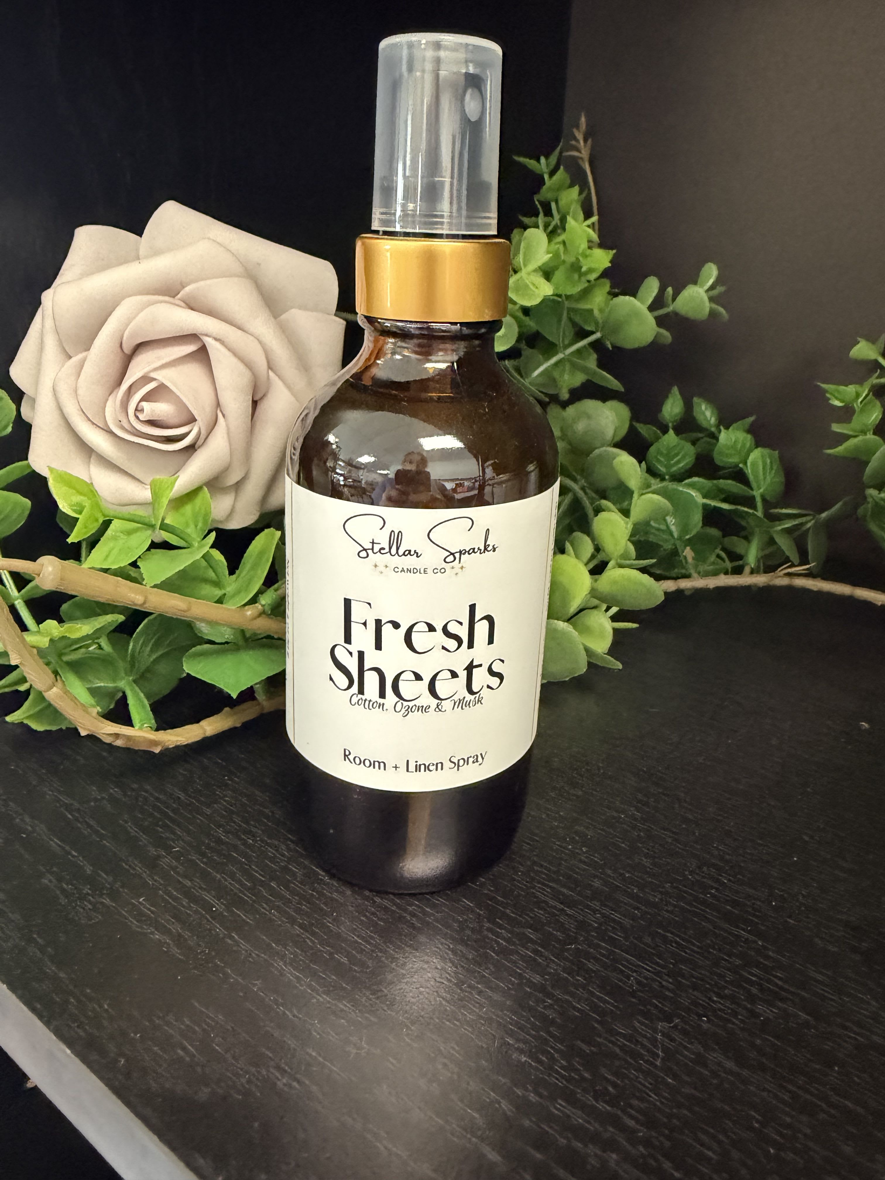 Fresh Sheets Room Spray - The Fresh Sheets room and linen spray is a giftable home fragrance that feels like freshly made beds and quiet mornings - perfect as a housewarming present, a host gift, or a thoughtful "thinking of you" gesture. This room spray product pairs an amber glass bottle labeled "Fresh Sheets" with a clear cap and gold collar, presented with a soft beige rose and bright green faux foliage for a spa-like, neutral look that reads as both elegant and approachable.  The bottle comes with a functional spray nozzle and is styled in a decorative basket for easy gifting and display. Visual contrast of white, green and warm amber tones gives it a calming, clean appearance that complements contemporary and classic interiors alike. While the rose and greenery are decorative accents rather than fresh blooms, their texture and color help convey the linen-fresh scent and make the package feel special.  Give this to someone who appreciates simple luxuries - a new homeowner, a hostess, or the person who loves to keep their space serene. Sized for easy placement on a bedside table or bathroom shelf, the spray is ready to use straight from the bottle; store out of direct sunlight to keep the packaging and scent looking and smelling their best.
