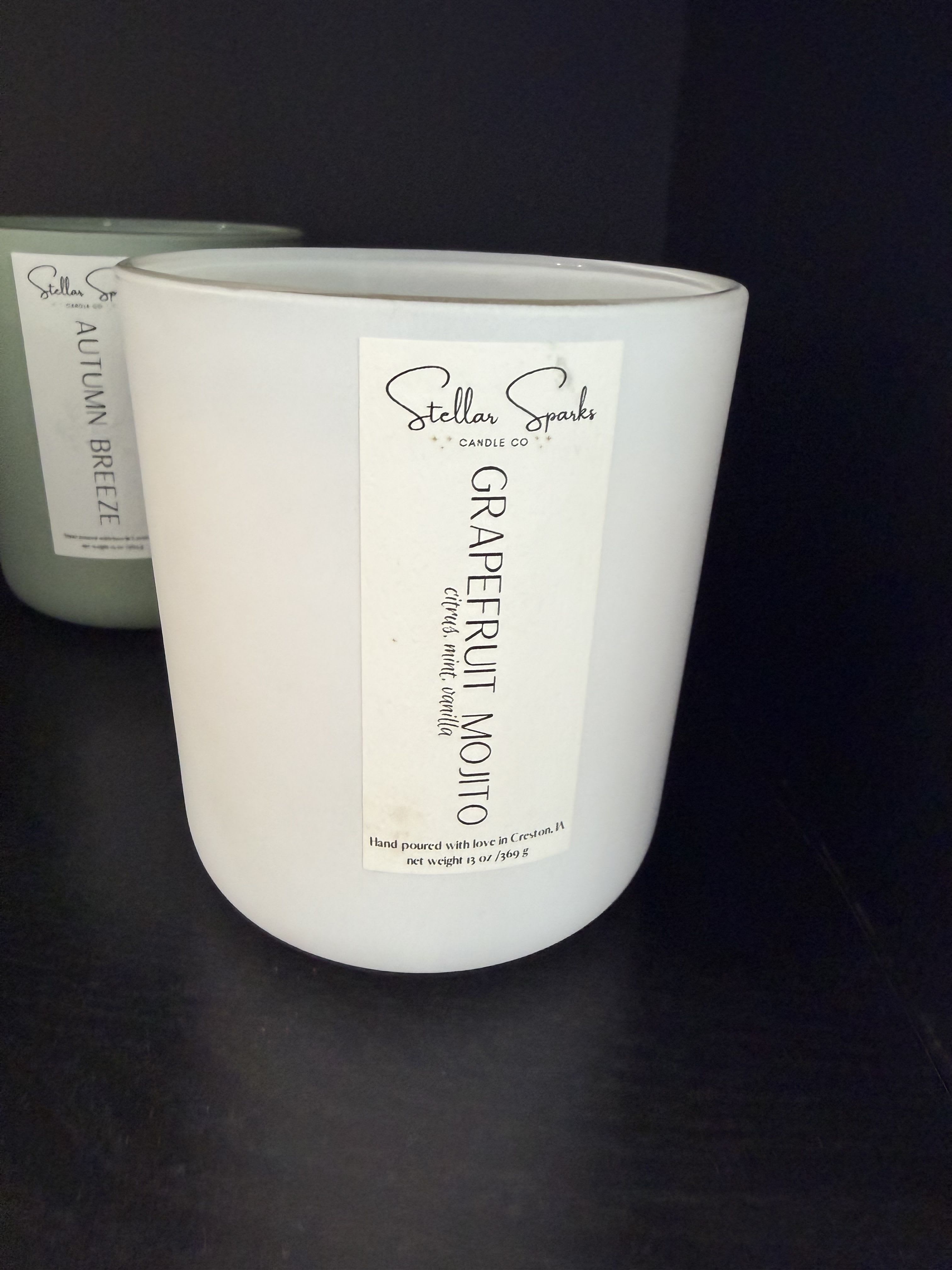 Grapefruit Mojito 13oz Candle - Bright, citrusy top notes meet cool mint in a home fragrance gift that feels like a sunlit kitchen-introducing a Grapefruit Mojito 13oz candle perfect for housewarming gifts or everyday moments of refreshment. As a scented candle for gifting, it creates an uplifting, clean atmosphere the person you're thinking of can enjoy while relaxing, cooking, or entertaining.  The 13oz candle comes in a simple cylindrical jar with a minimalist label-clean white wax in one jar and a muted sage-green companion visible behind it-paired with neutral black and green accents that suit modern or Scandinavian decor. The understated packaging and crisp typography make it easy to place on a mantel, bedside table, or kitchen counter without clashing with the room.  Give this as a thoughtful housewarming, hostess, or "thinking of you" present that says freshness and calm. For best results, trim the wick to 1/4" before each burn and allow the wax to melt to the jar edges on the first burn to ensure an even melt pool; the 13oz size offers many hours of fragrance for everyday enjoyment.